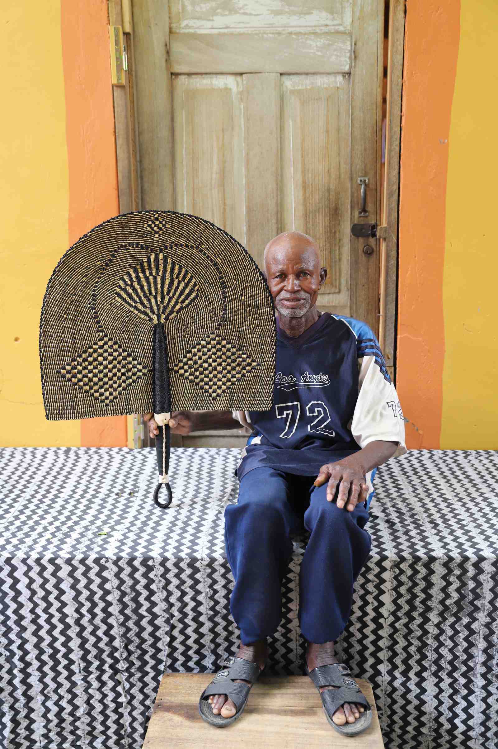 Naba Penga wall fan in natural straw with a dark geometric diamond weave, held by a seated person for scale.
