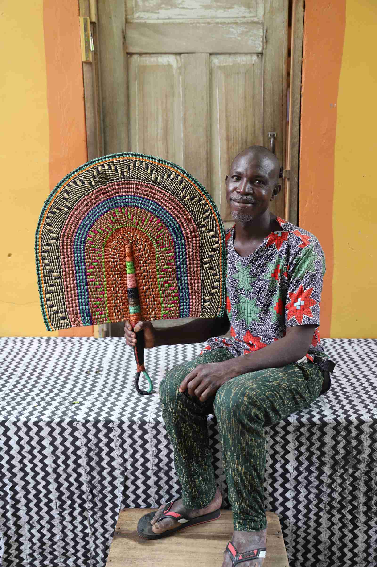 Naba Penga wall fan in natural straw with teal, rust and green multicolour bands, held by a seated person for scale.