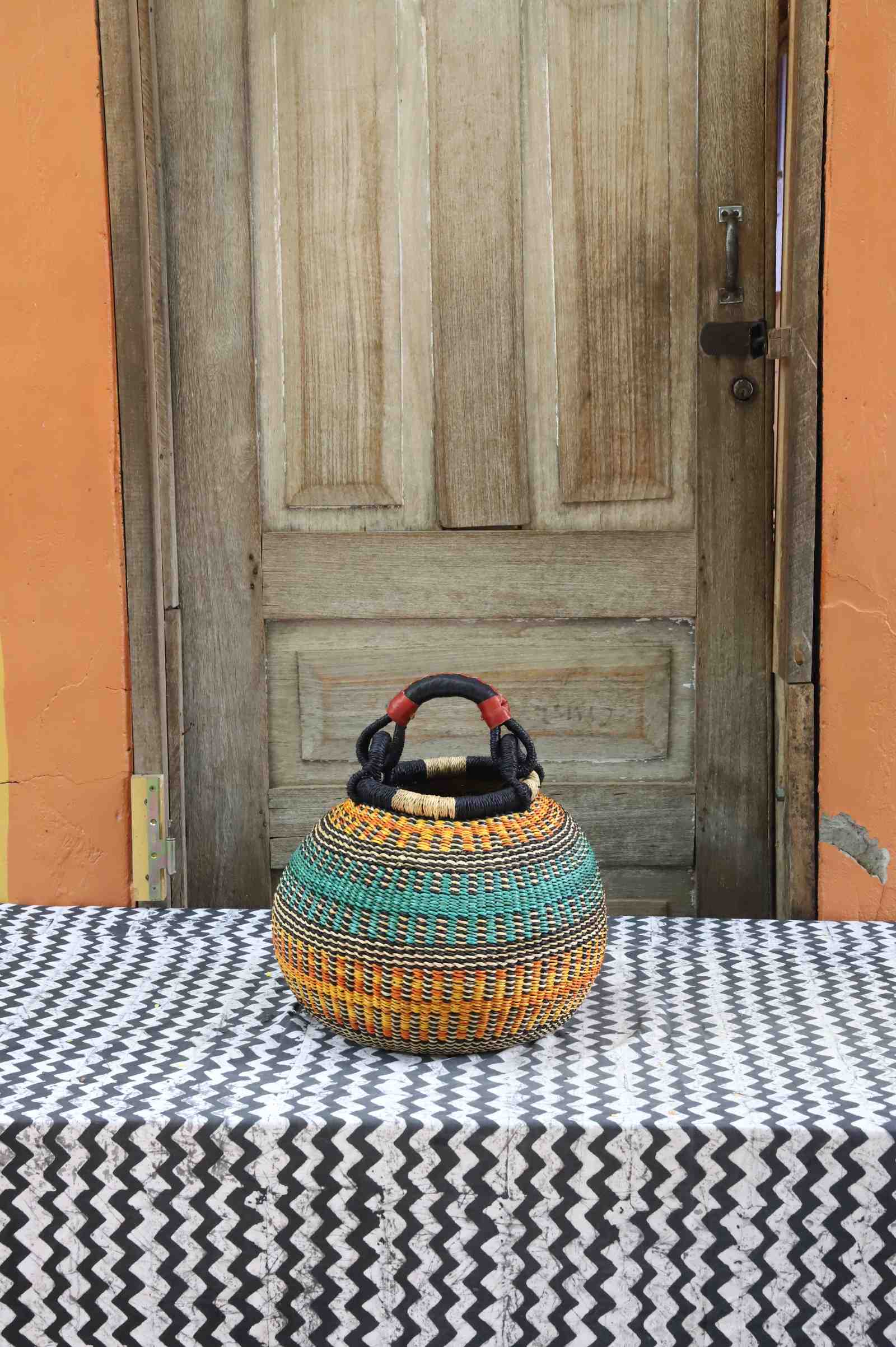 Handwoven Bolga pot basket in natural straw with orange and teal woven bands and twin leather-wrapped handles.