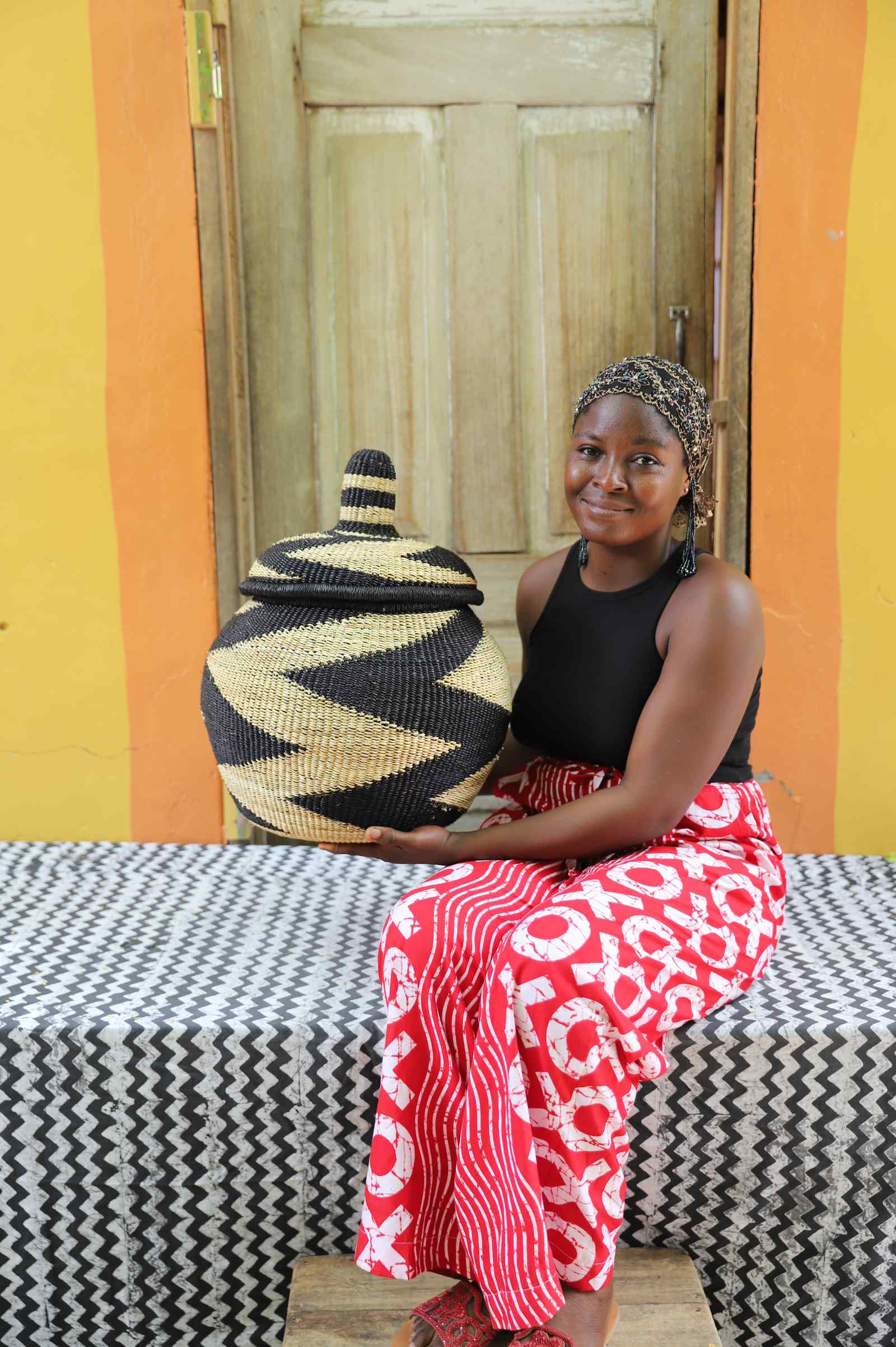 Lidded Bolga storage basket in a black and natural zigzag pattern, held by a seated woman for scale.