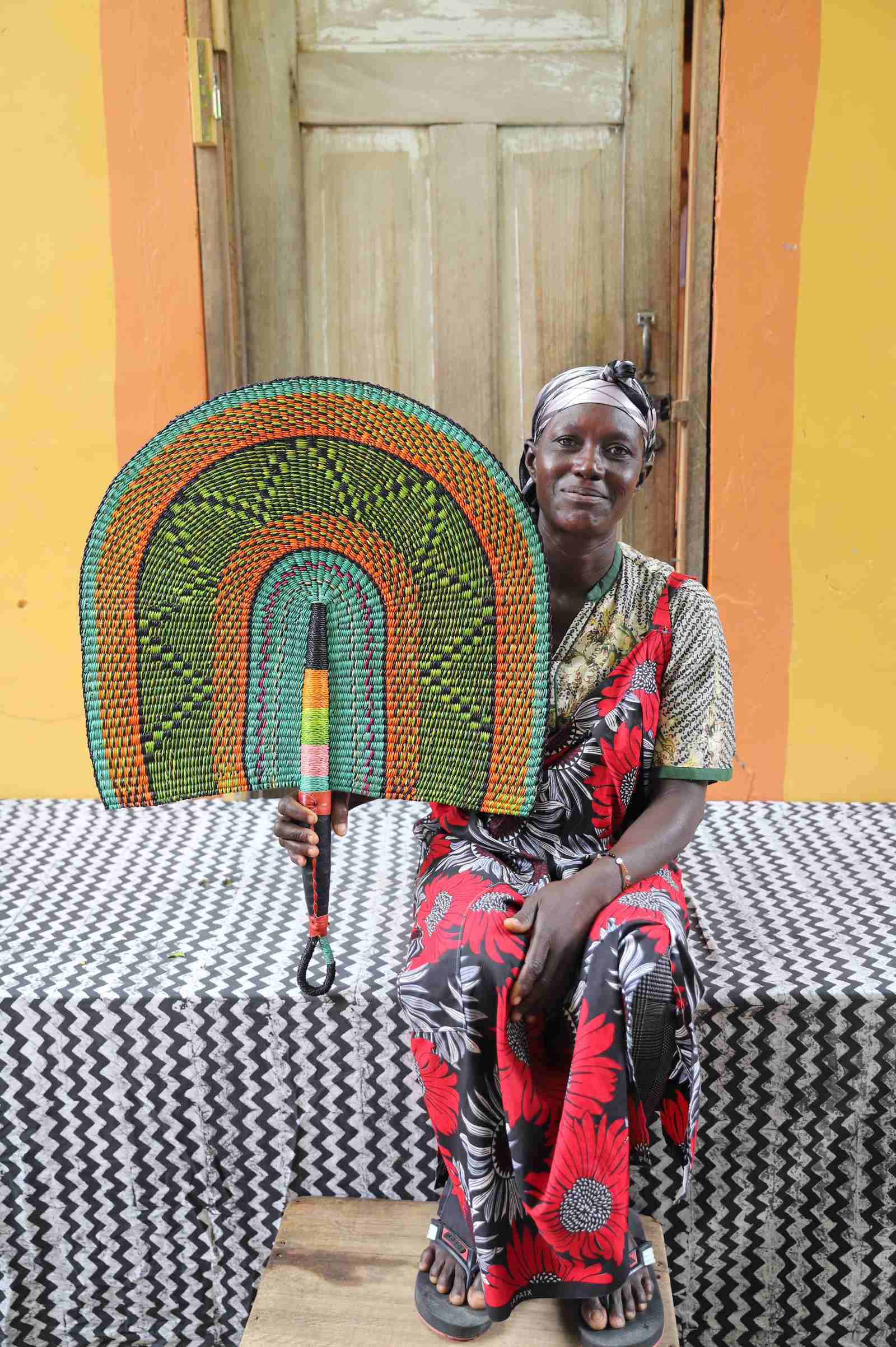 Naba Penga wall fan in green woven straw with an orange border, held by a seated person for scale.