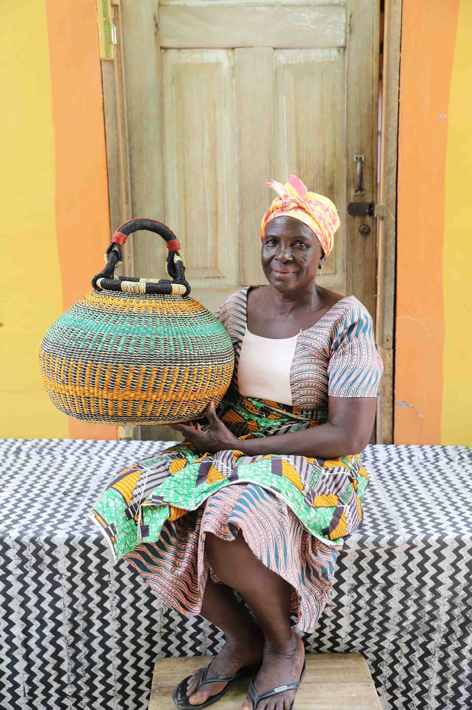 Round Bolga pot basket in orange and green stripes with a leather-wrapped handle, held by a seated woman.