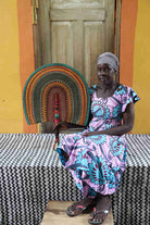 Naba Penga wall fan in natural straw with teal, orange and green concentric bands, held by a seated person for scale.