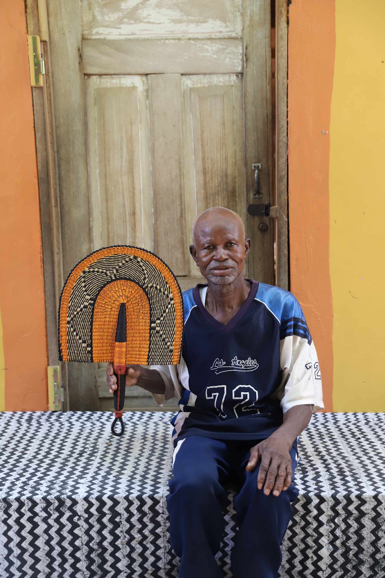 Weaver holding a handwoven Bolga Fan in natural straw with black geometric outer arch, in Bolgatanga, Ghana.