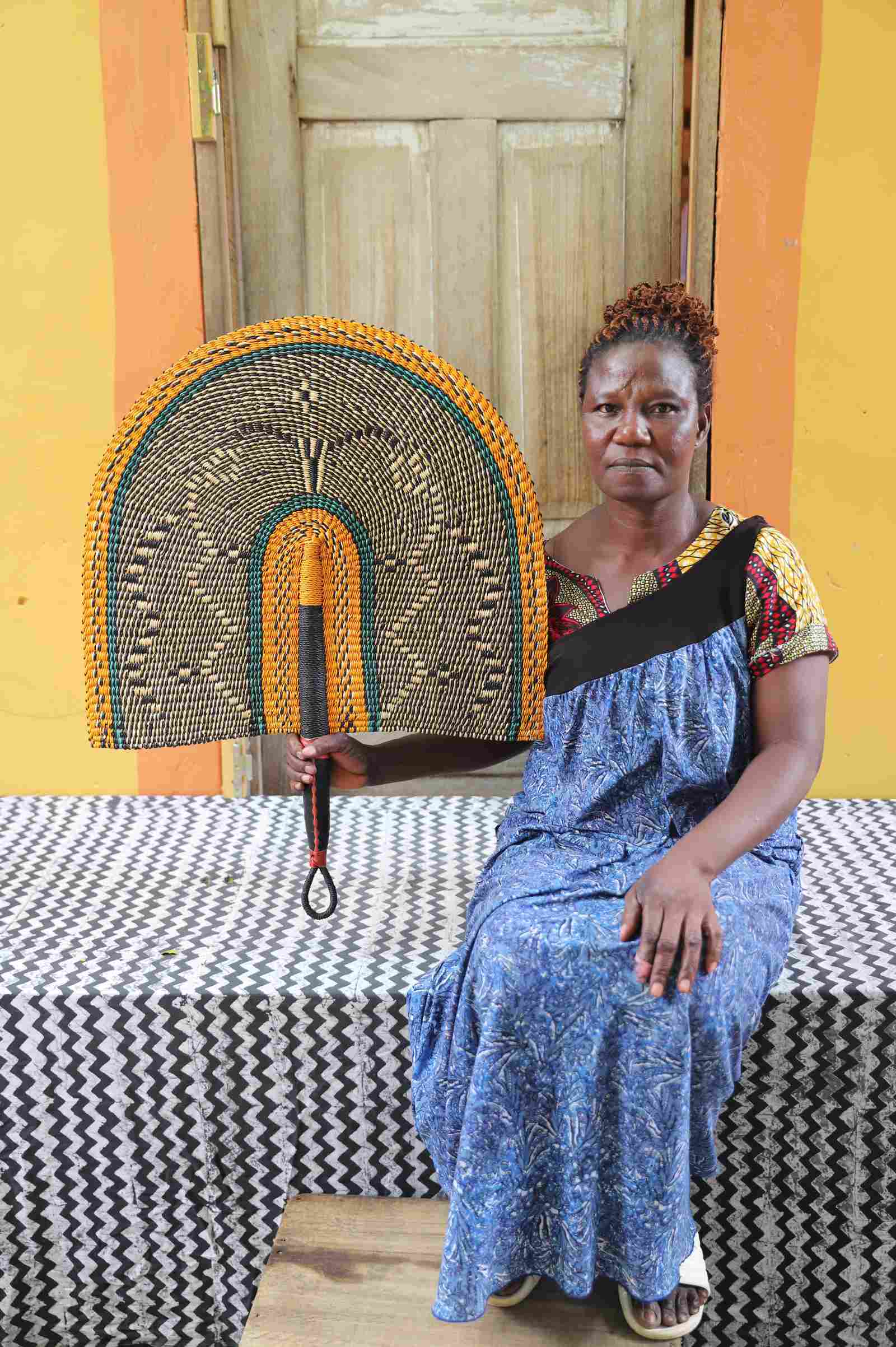Naba Penga wall fan in natural straw with a mustard border and green trim, held by a seated person for scale.