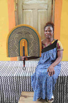 Naba Penga wall fan in natural straw with a mustard border and green trim, held by a seated person for scale.