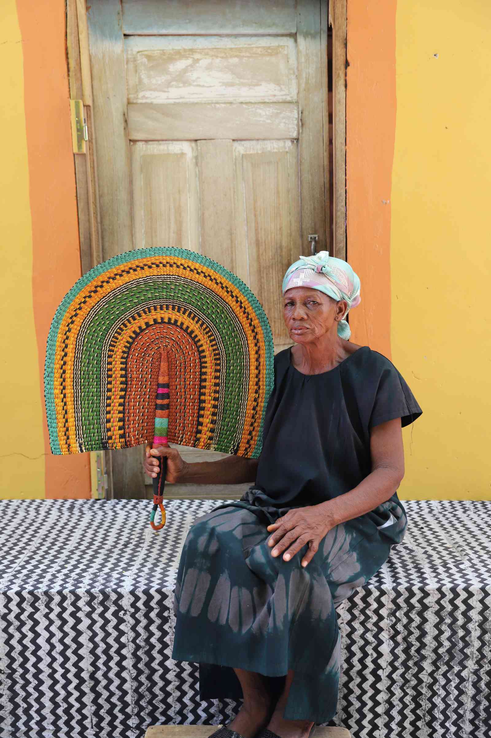 Weaver holding a Naba Penga handwoven wall fan with arch bands in green, amber and terracotta, in Bolgatanga, Ghana.