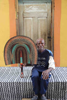 Naba Penga wall fan in natural straw with a teal inner arch and dark patterned border, held by a seated person for scale.