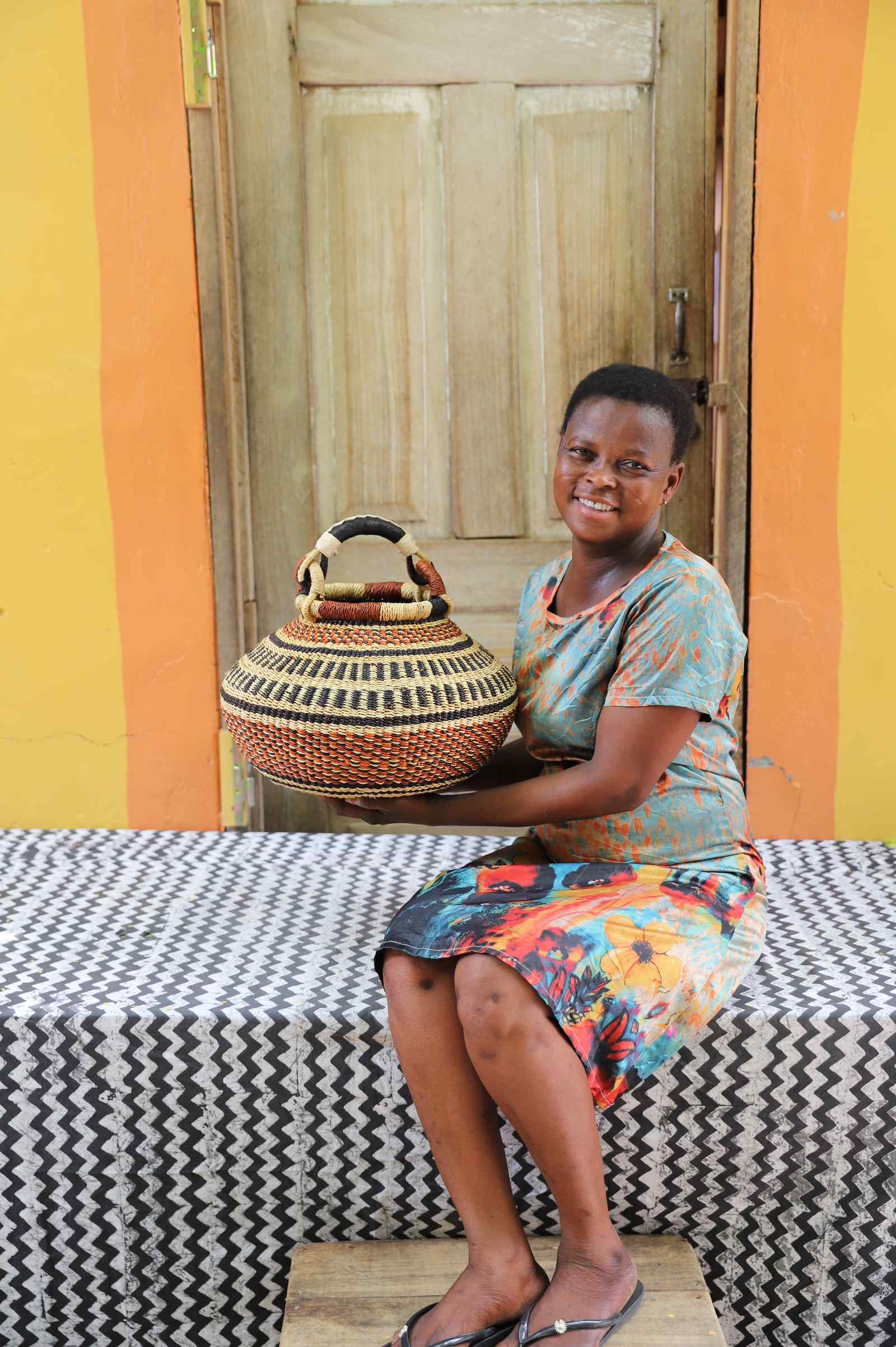 Small round Bolga pot basket in black and cream diamond weave with a rust band, held by a seated woman for scale.