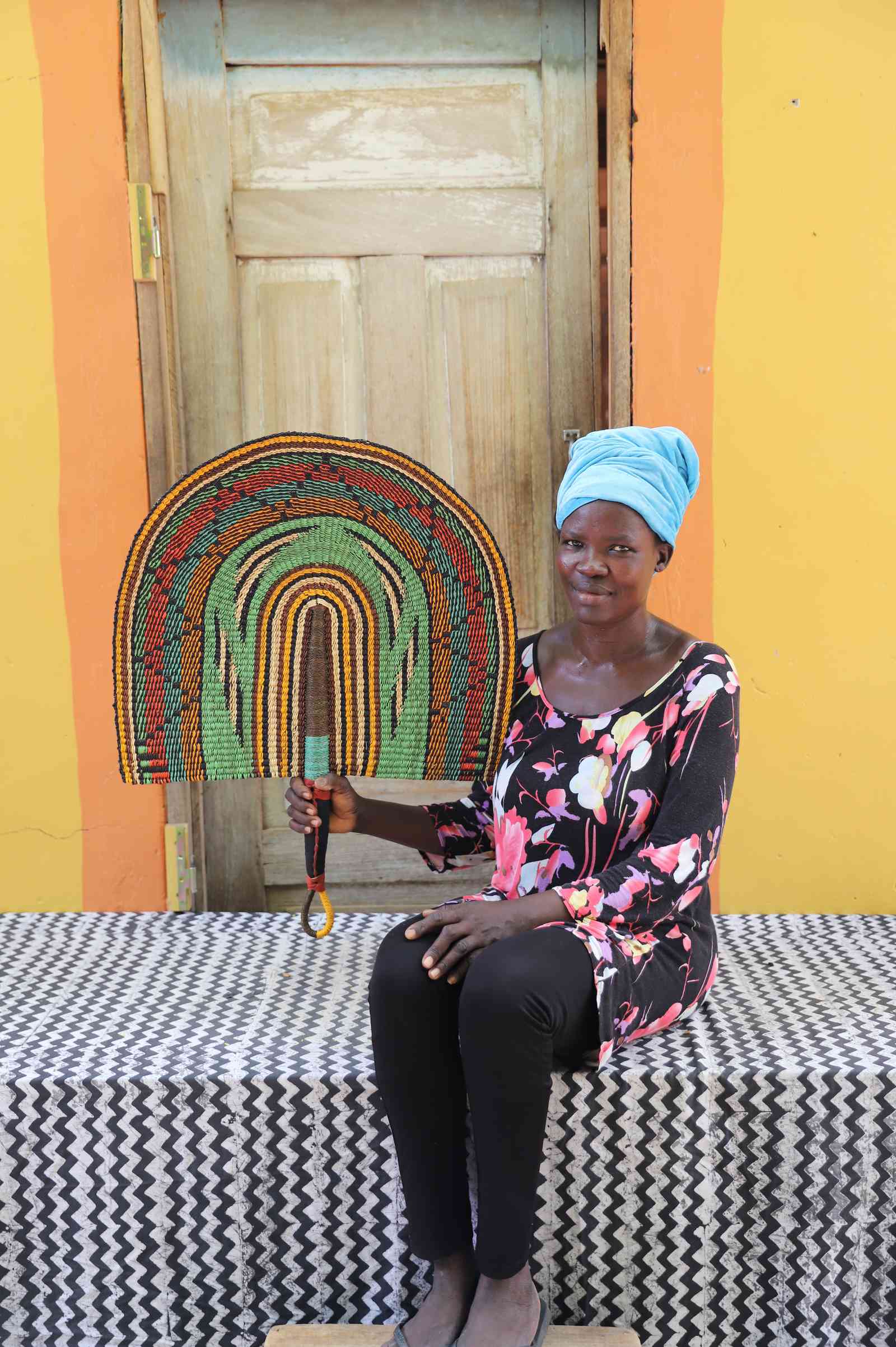 Weaver holding a Naba Penga handwoven wall fan with arch bands in green, terracotta and amber, in Bolgatanga, Ghana.
