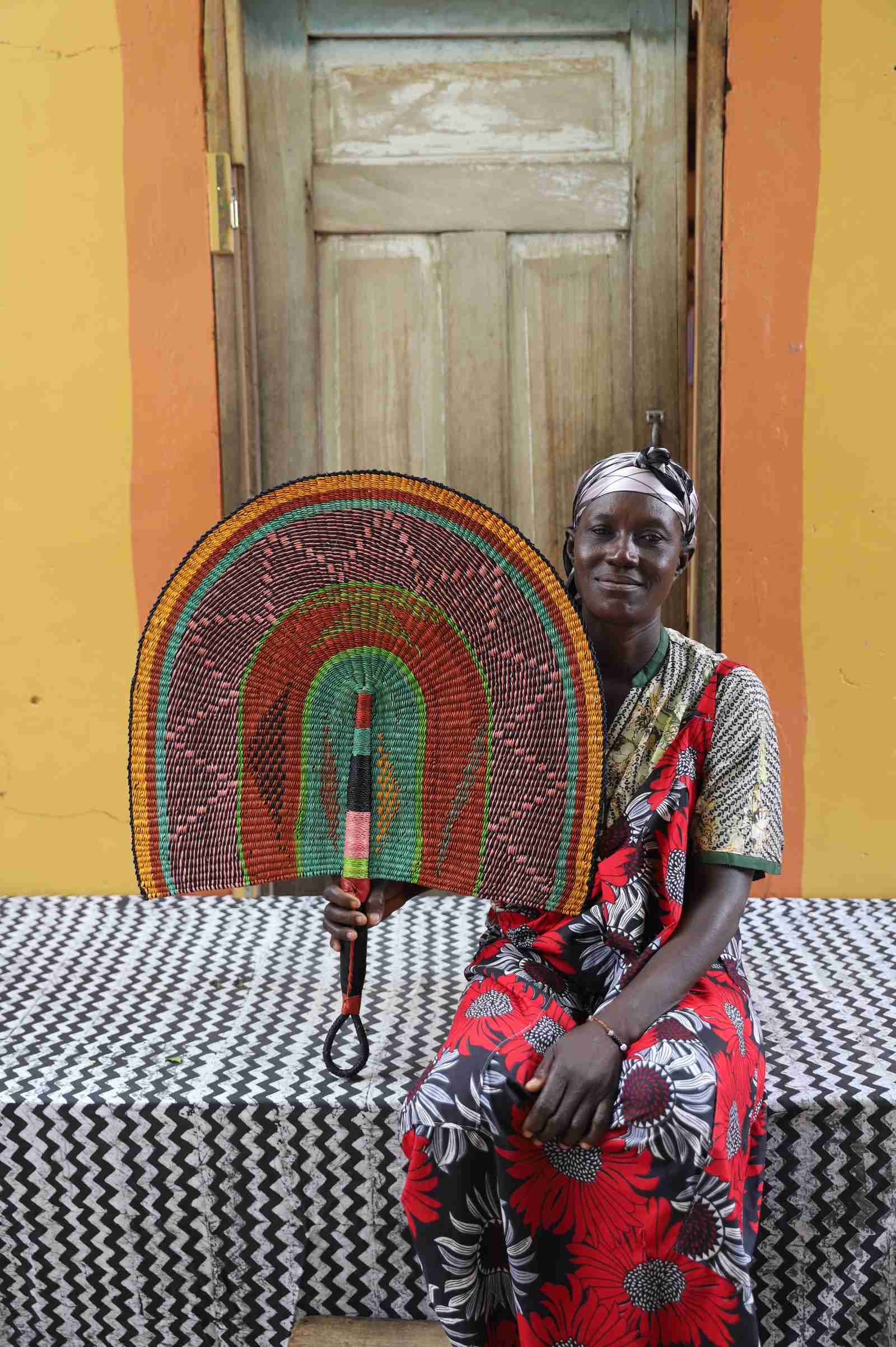 Naba Penga wall fan in natural straw with green, orange and black concentric bands, held by a seated person for scale.