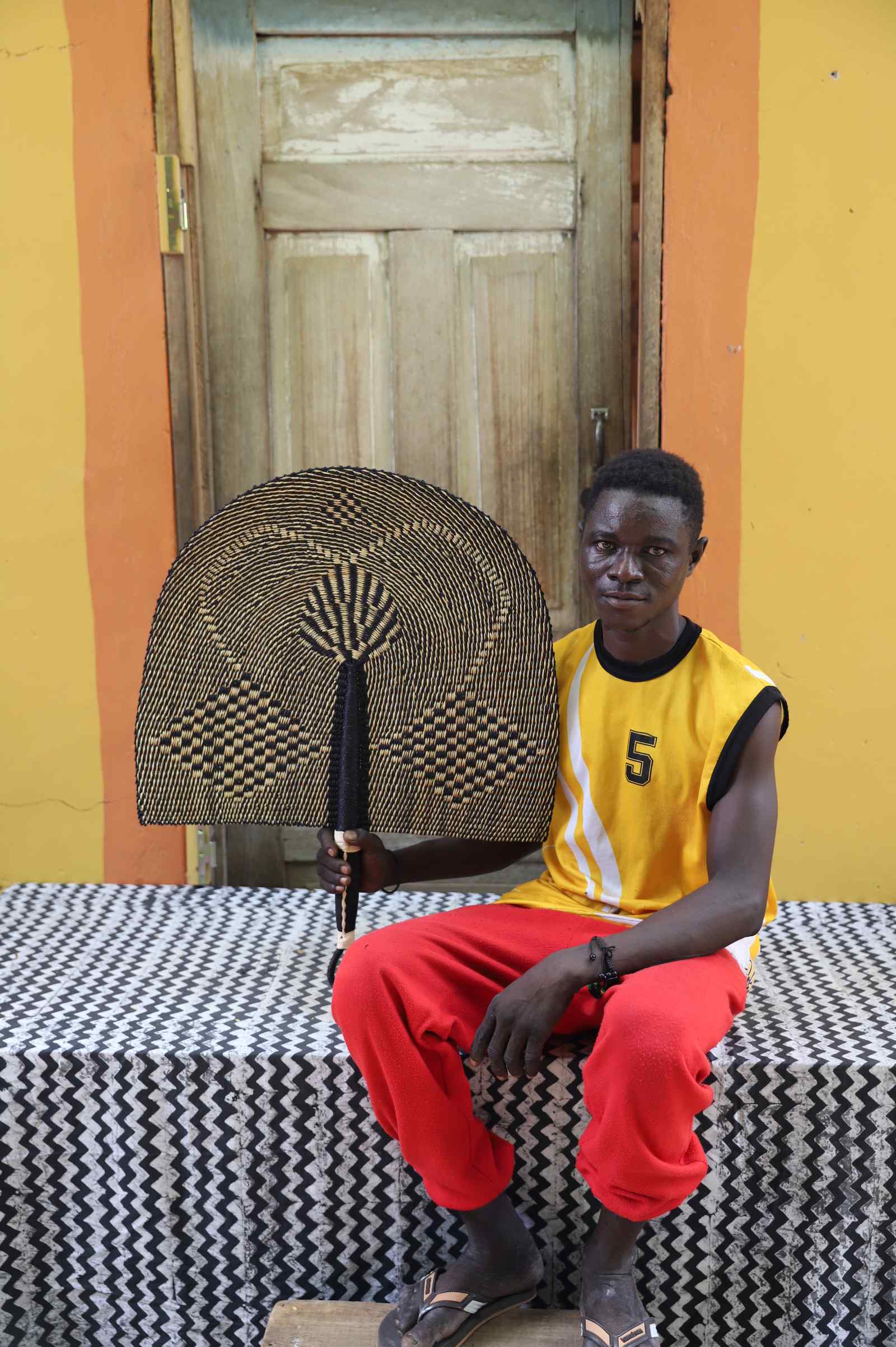 Weaver holding a Naba Penga handwoven wall fan in natural straw and black geometric check, in Bolgatanga, Ghana.