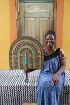 Naba Penga wall fan in natural straw with rust, green and teal concentric arches, held by a seated person for scale.