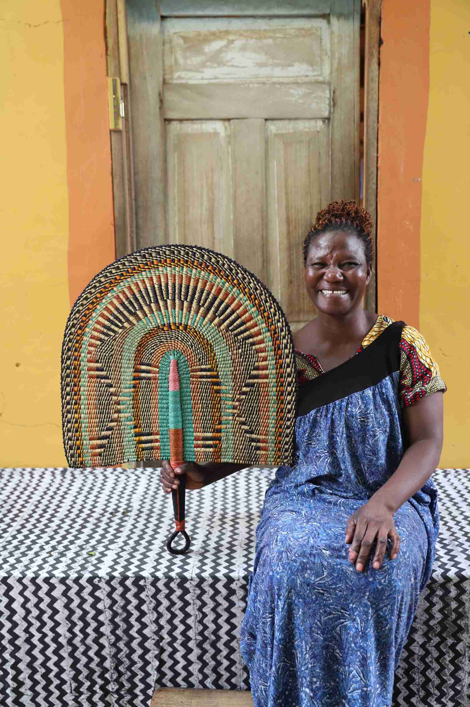 Naba Penga wall fan in natural straw with rust, green and teal concentric arches, held by a seated person for scale.