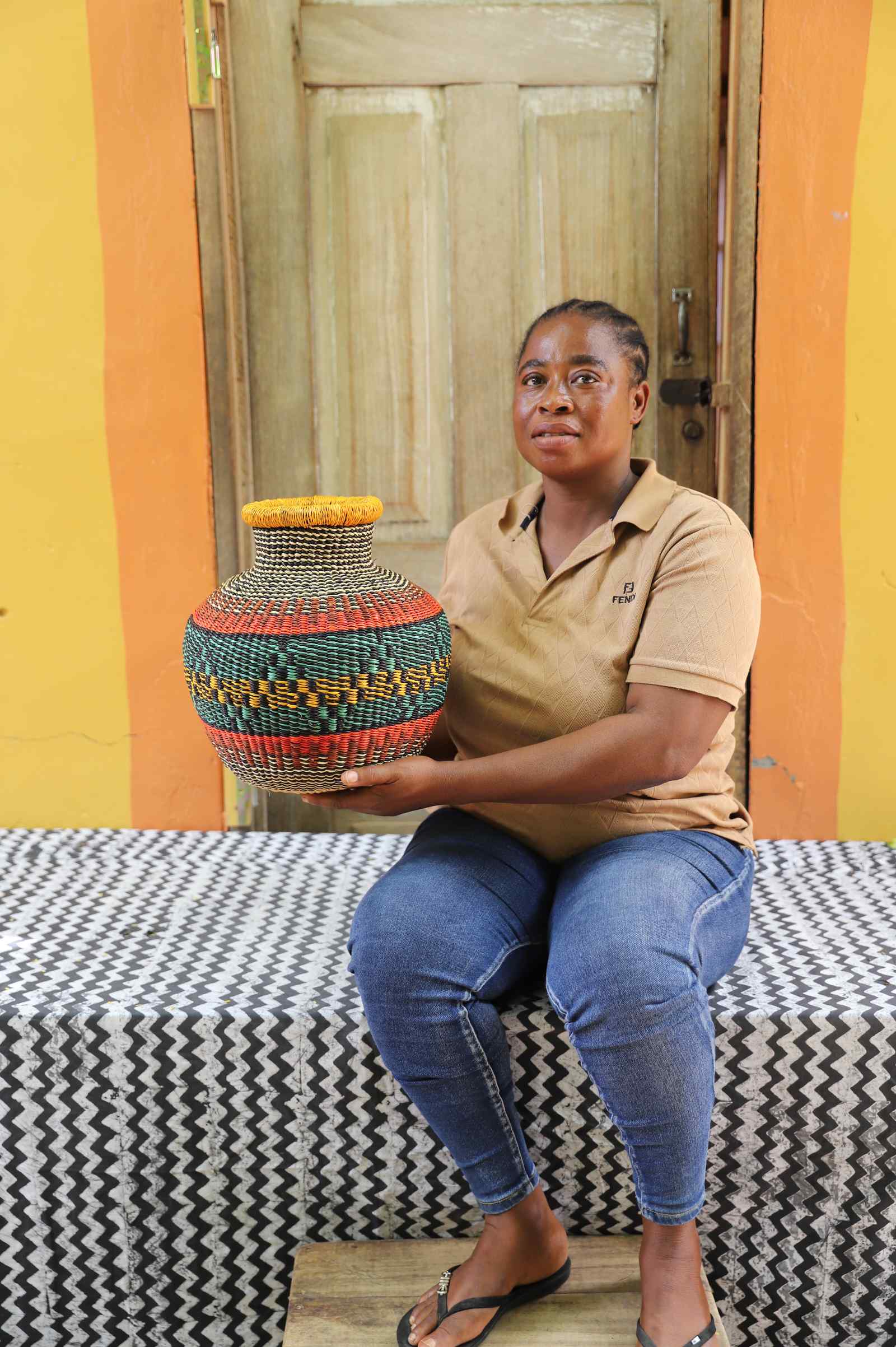 Small round Bolga pot basket in teal, orange and yellow diamond weave, held by a seated woman for scale.