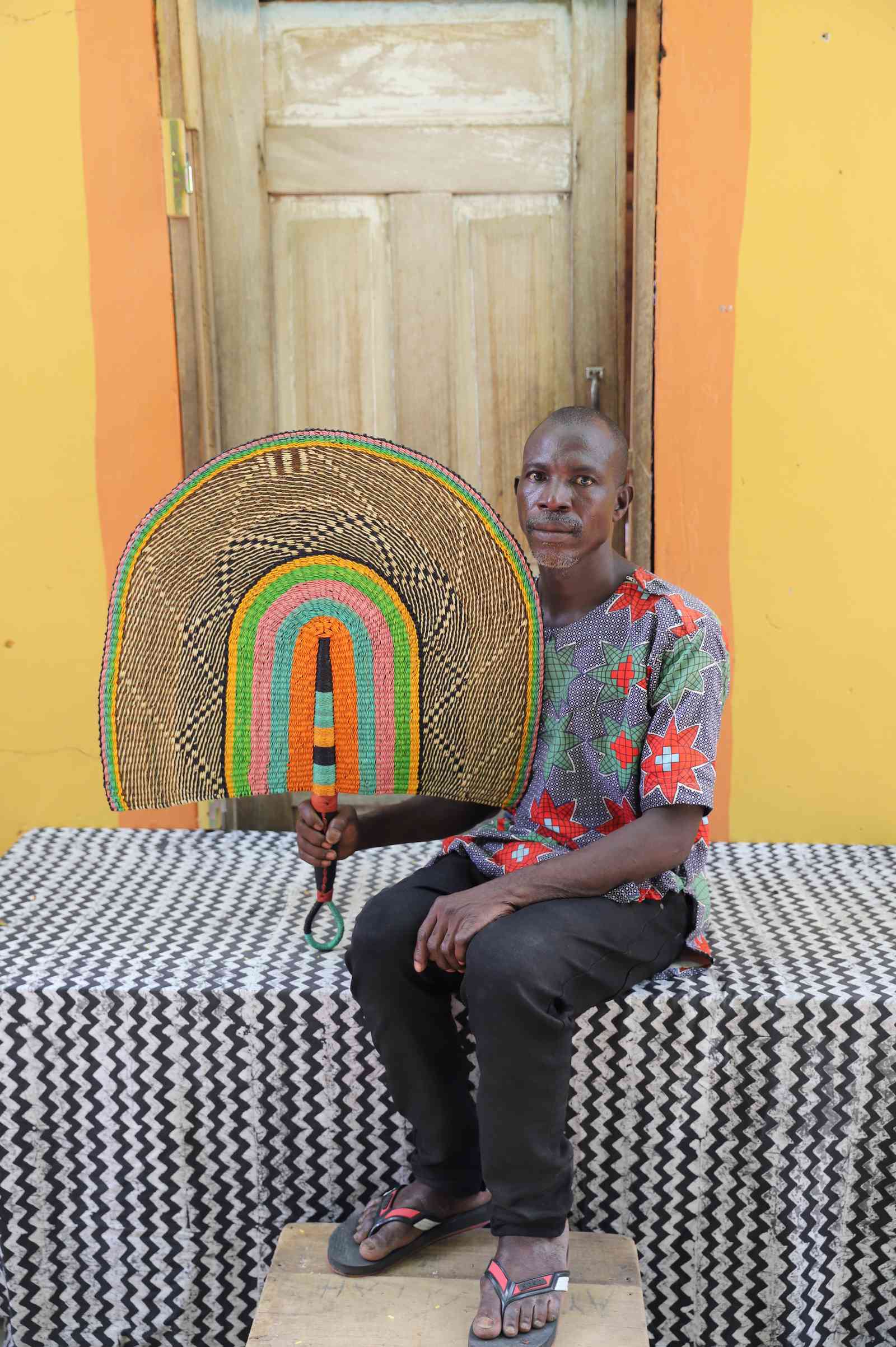 Weaver holding a Naba Penga handwoven wall fan in natural straw with pink, green and orange inner arches, in Bolgatanga.
