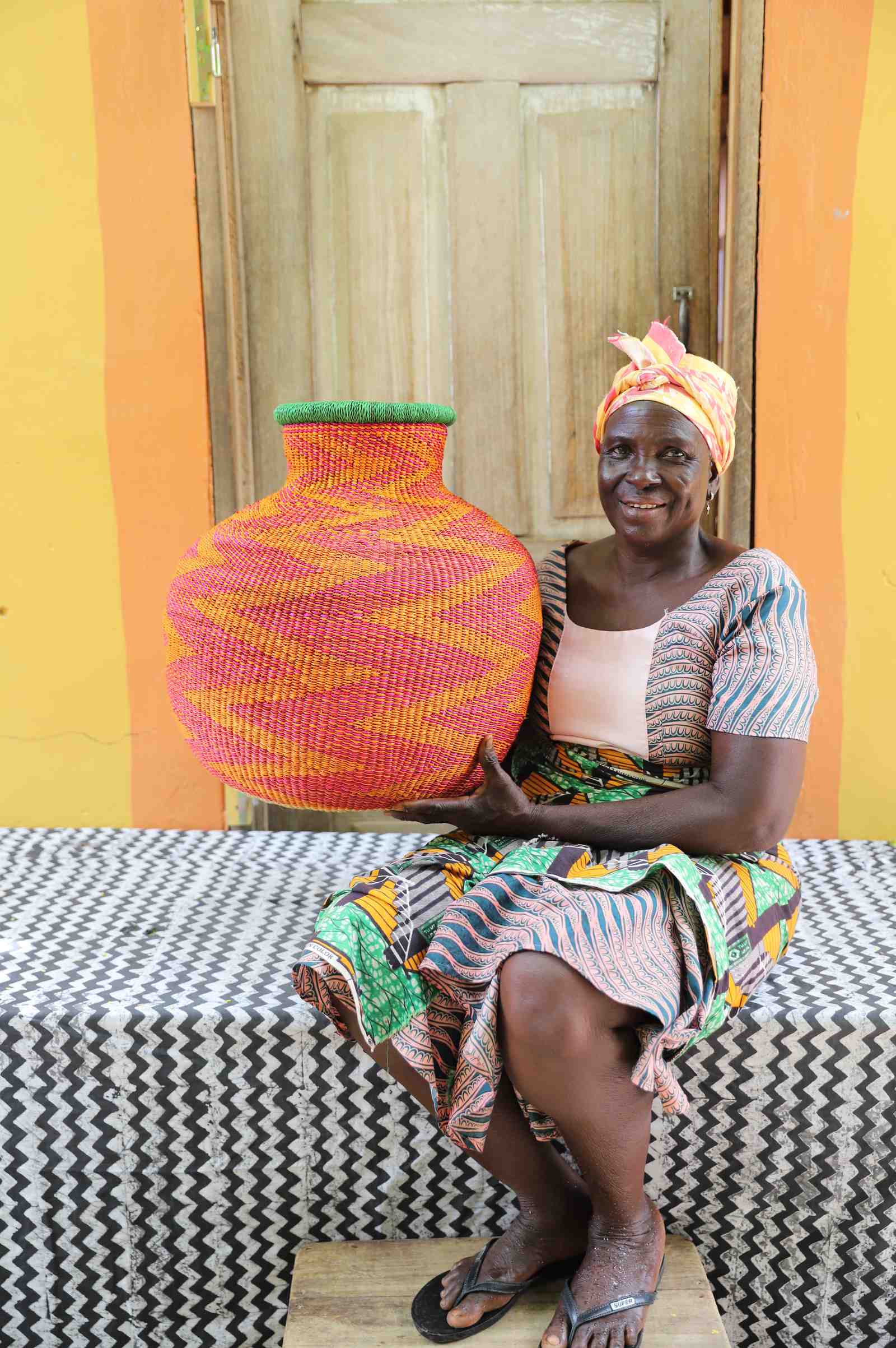 Large round Bolga pot basket in pink and yellow zigzag weave with a green rim, held by a seated woman for scale.