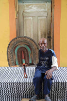 Naba Penga wall fan in natural straw with a teal arch and orange patterned border, held by a seated person for scale.
