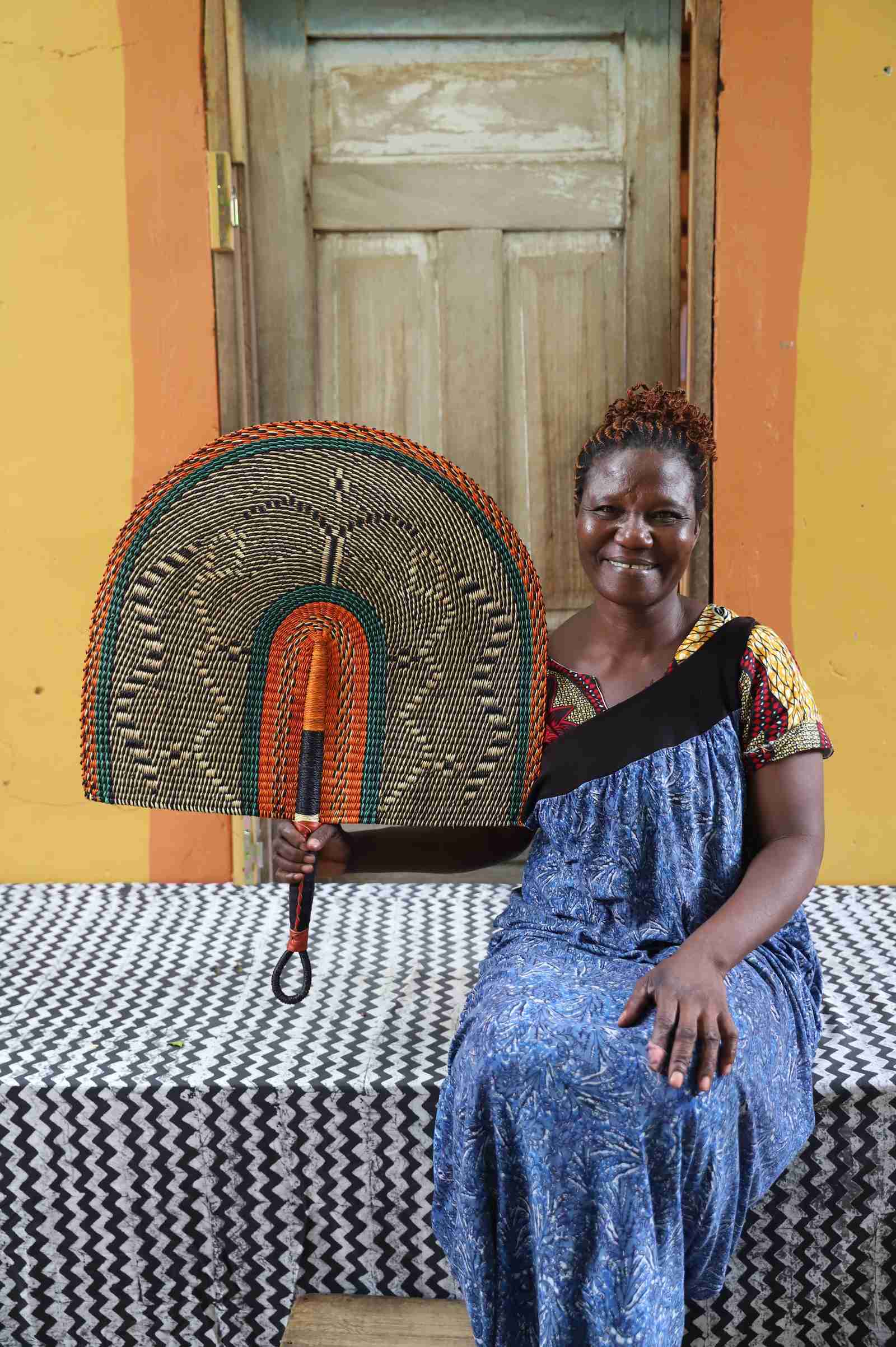 Naba Penga wall fan in natural straw with an orange and green banded border, held by a seated person for scale.