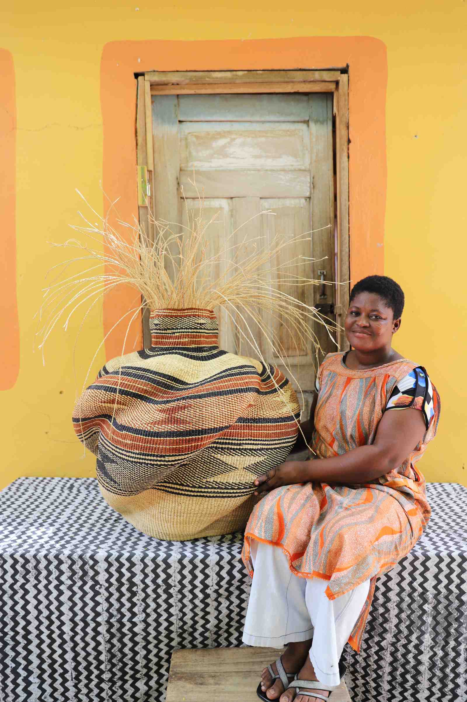 Round Bolga basket in tan with a rust and black striped band and loose straw fronds, shown beside a seated woman for scale.