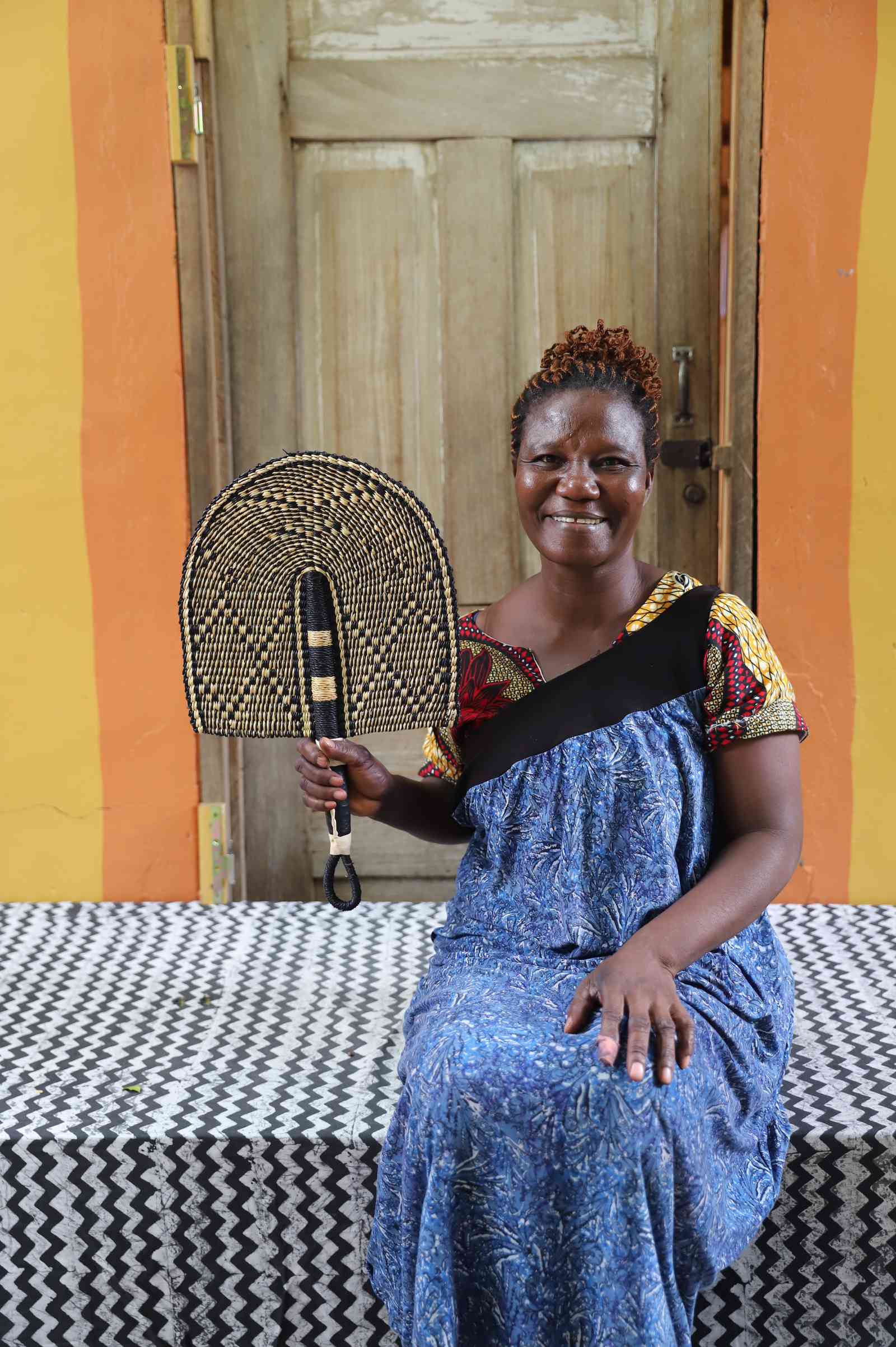 Bolga hand fan in natural straw with a dark chevron pattern, held by a seated person for scale.