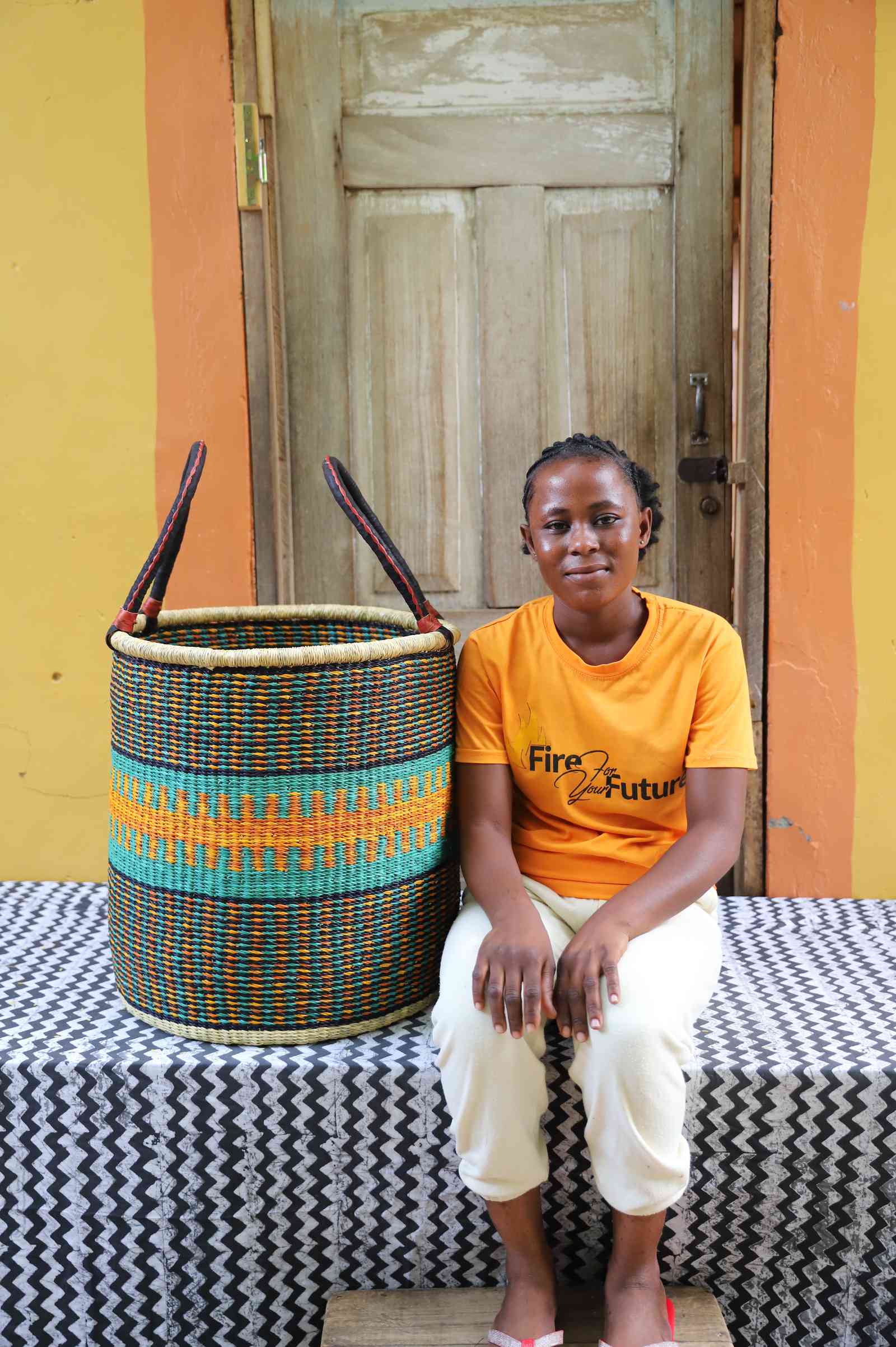 Tall open Bolga laundry basket with a teal and orange patterned band, shown beside a seated woman for scale.