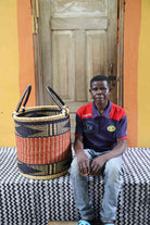 Open laundry basket in natural straw with an orange and black zigzag band, shown beside a seated person for scale.