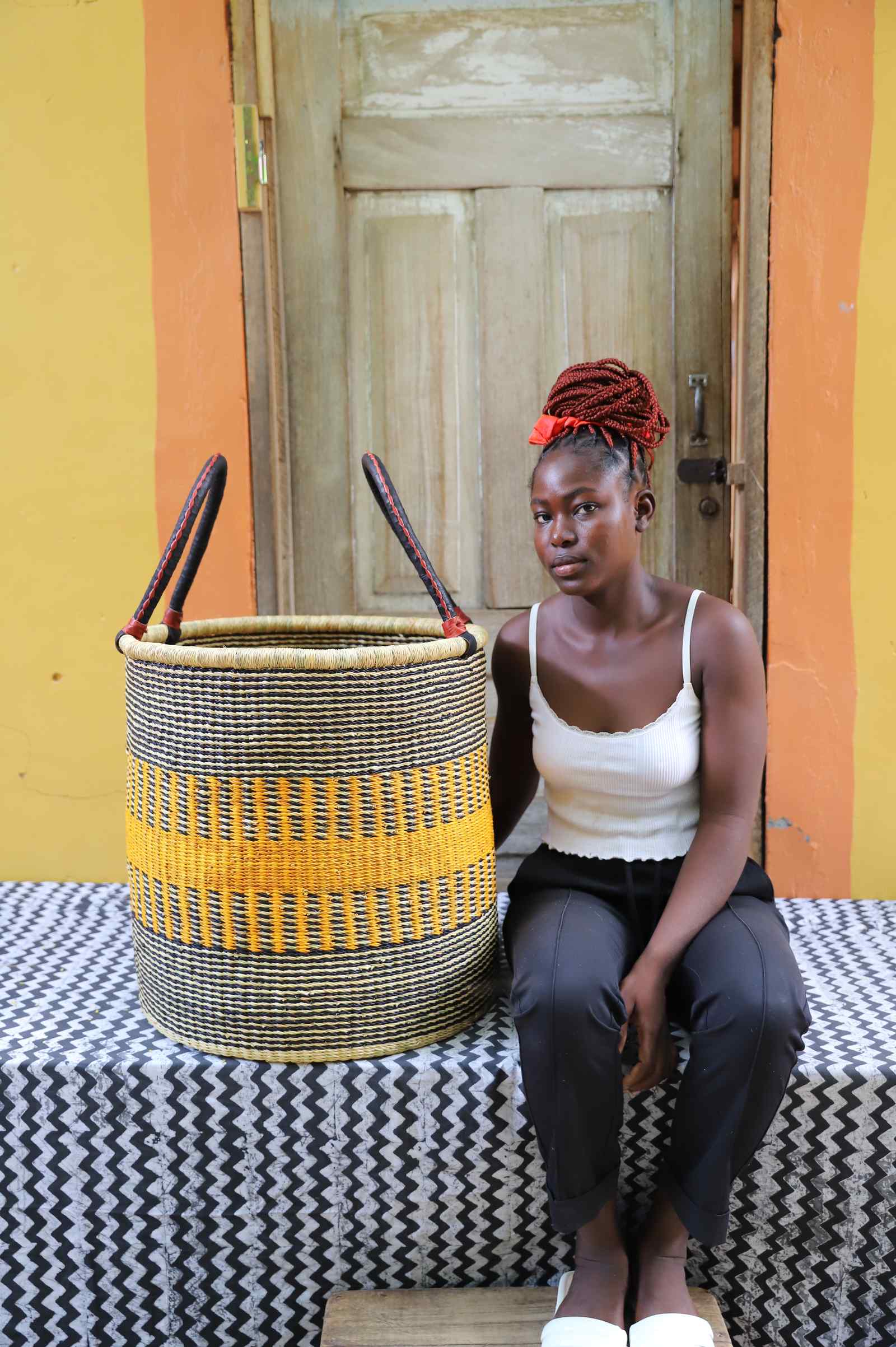 Tall open Bolga laundry basket with a bold yellow stripe band, shown beside a seated woman for scale.
