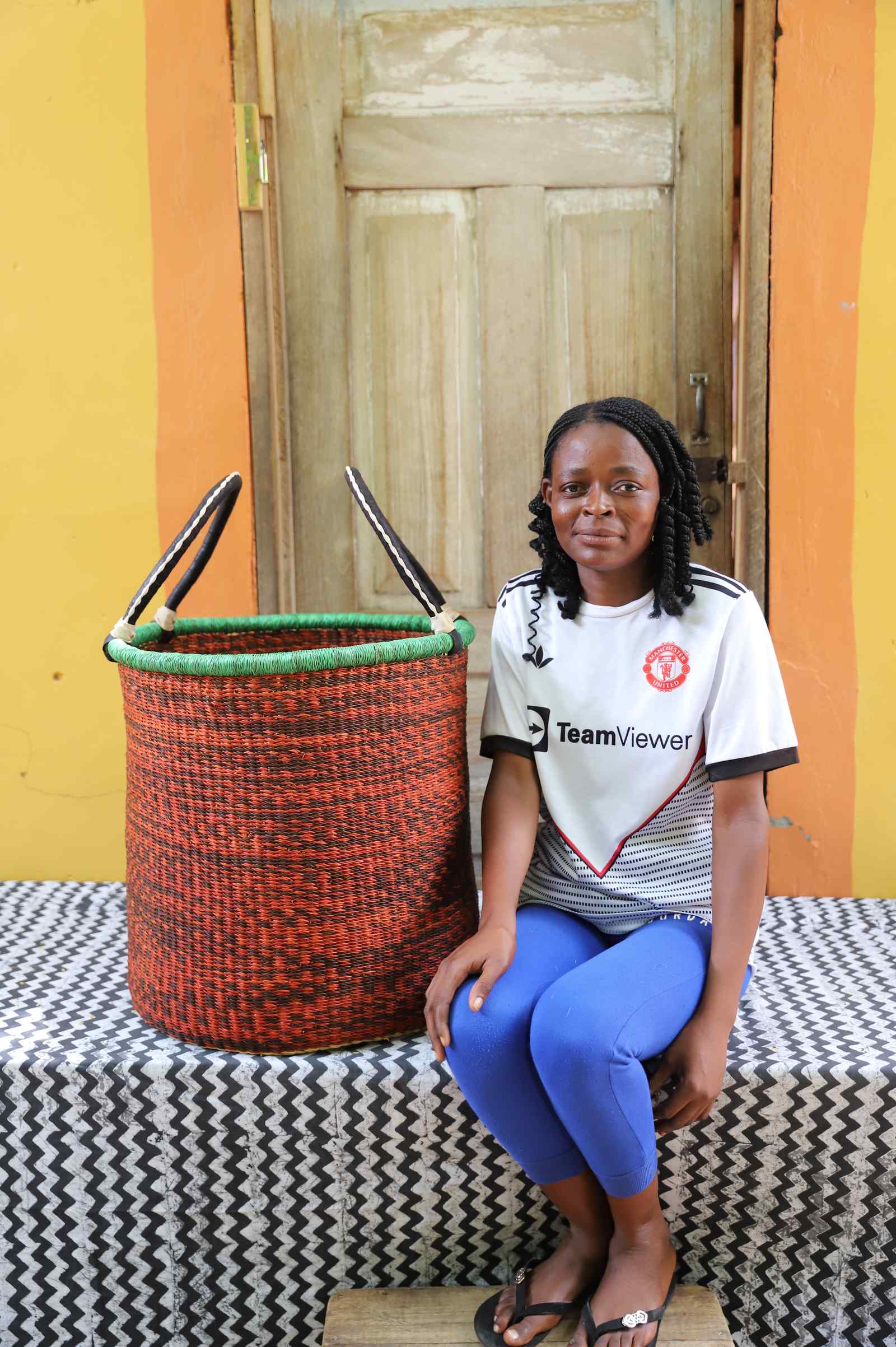 Tall open Bolga laundry basket in speckled rust-red weave, shown beside a seated woman for scale.