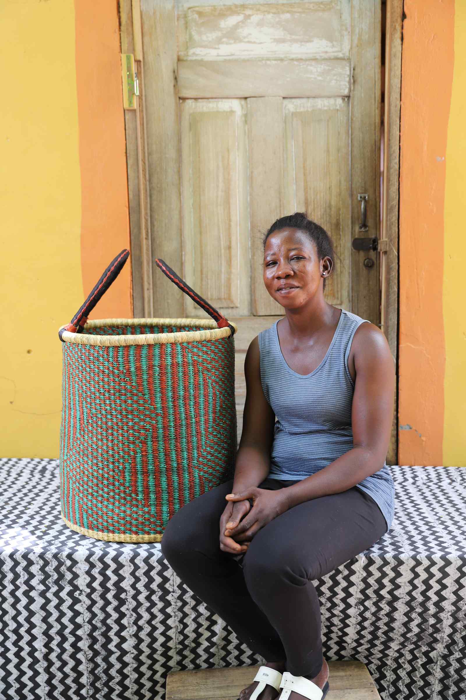 Tall open Bolga laundry basket in a green and rust striped weave, shown beside a seated woman for scale.