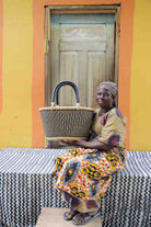 Large oval Bolga shopping basket in natural with dark zigzag, held by a seated woman in a printed dress.