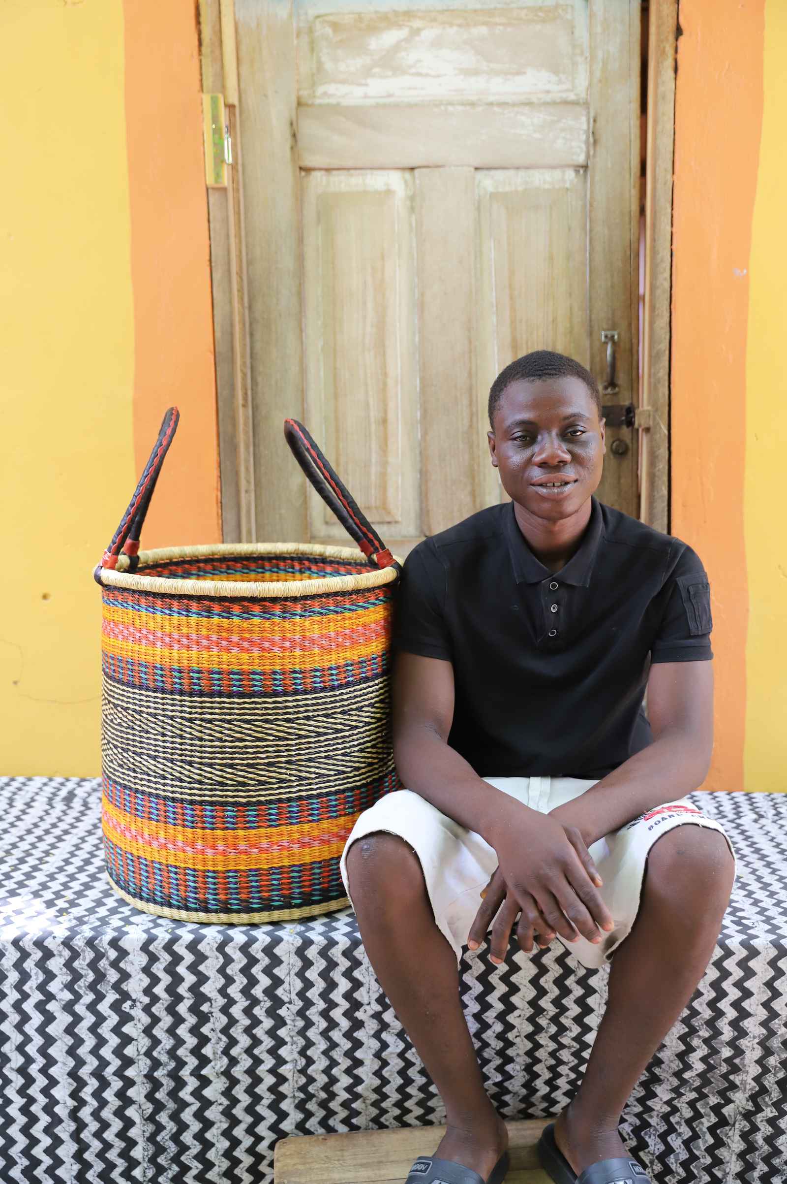 Tall open Bolga laundry basket with an orange, blue and cream striped band, shown beside a seated man for scale.