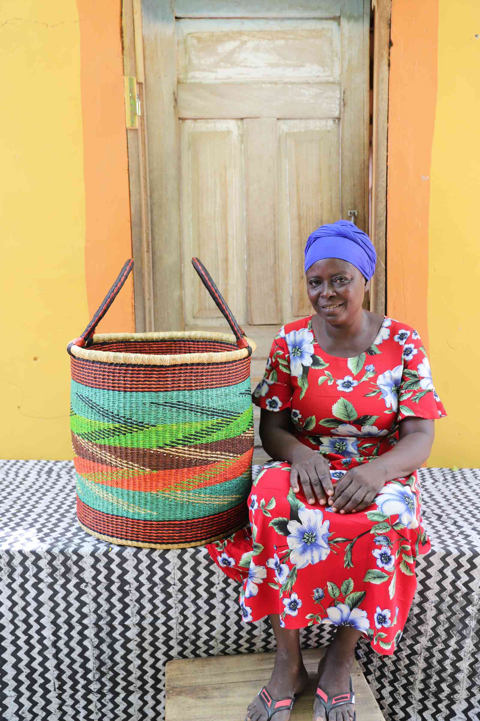Tall open Bolga laundry basket with a multicolour diagonal striped weave, shown beside a seated woman for scale.