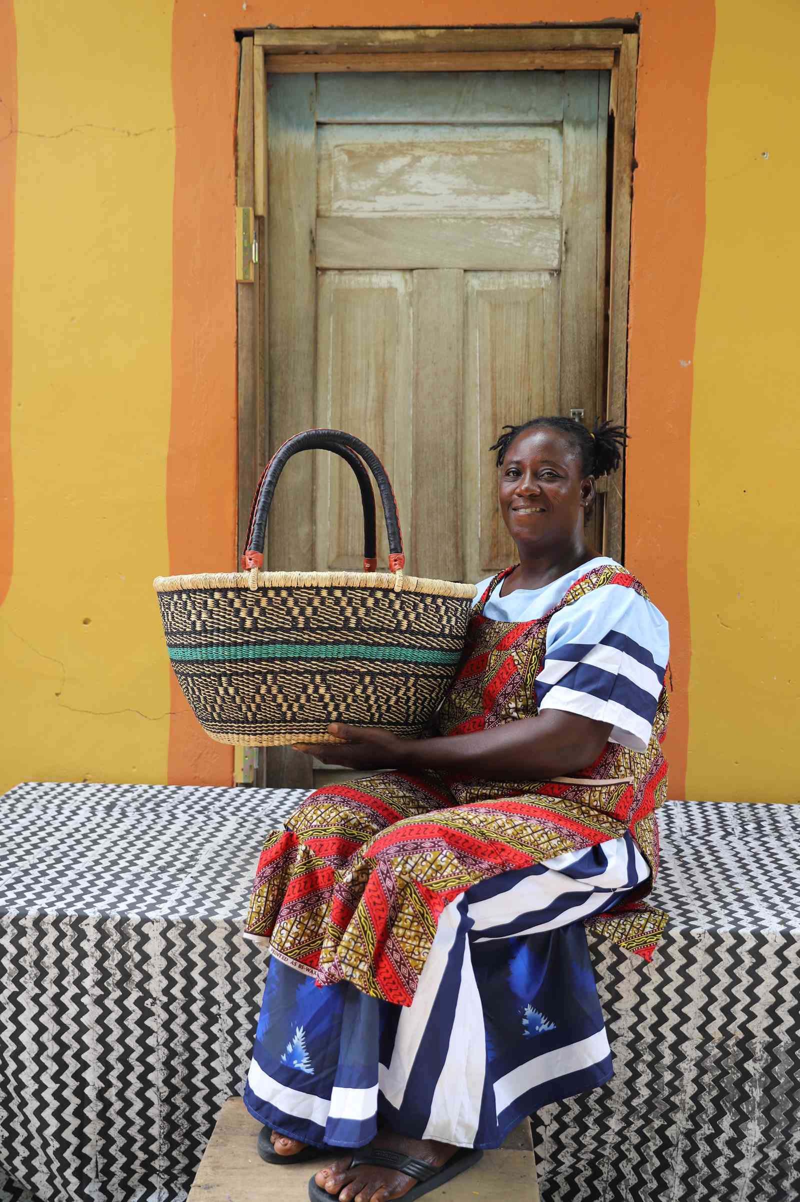 Large oval Bolga shopping basket in natural with teal and orange band, held by a seated woman in a printed dress.