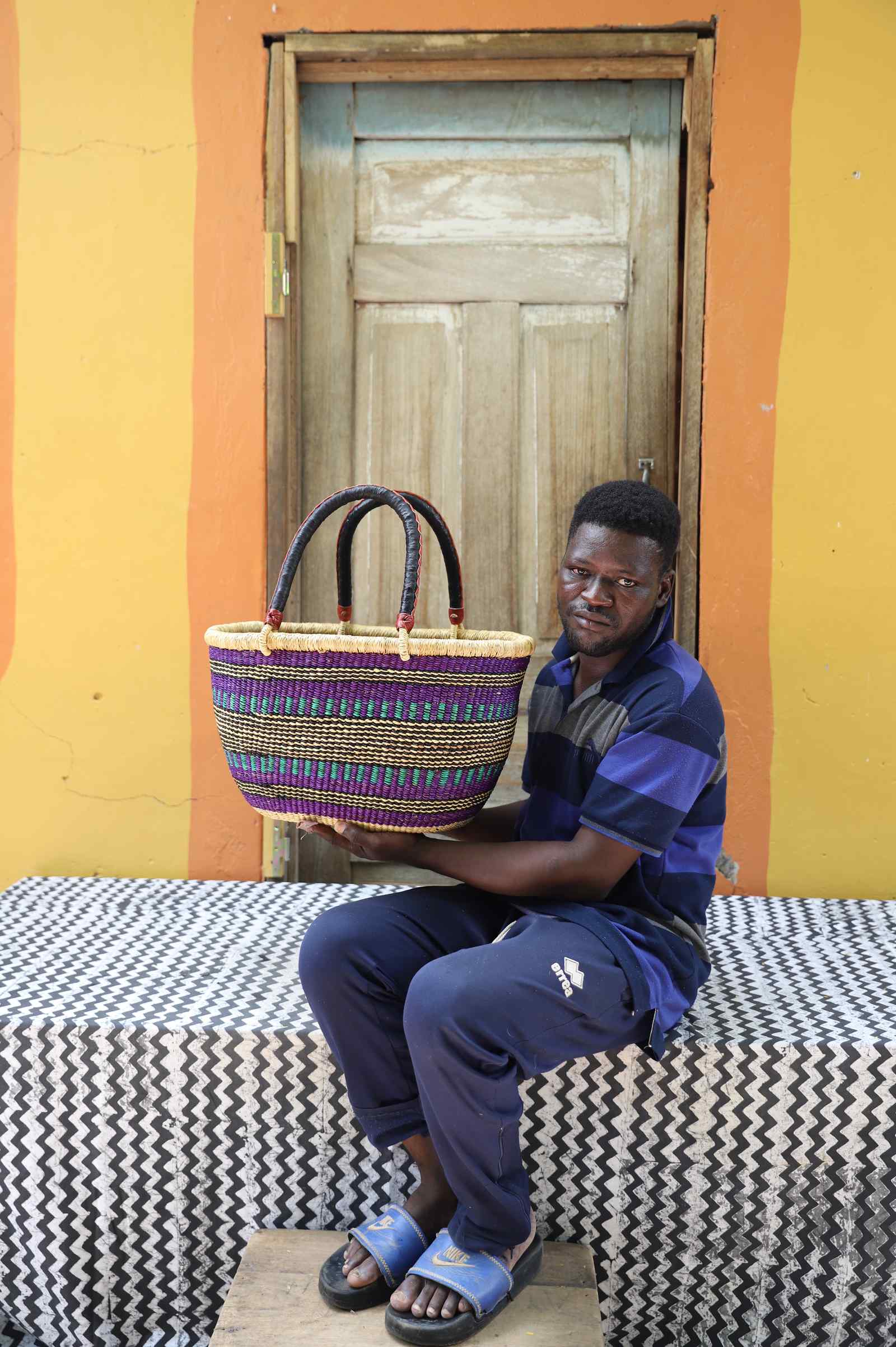 Medium oval Bolga shopping basket in fine multicolour stripes, held by a seated man in a dark top.