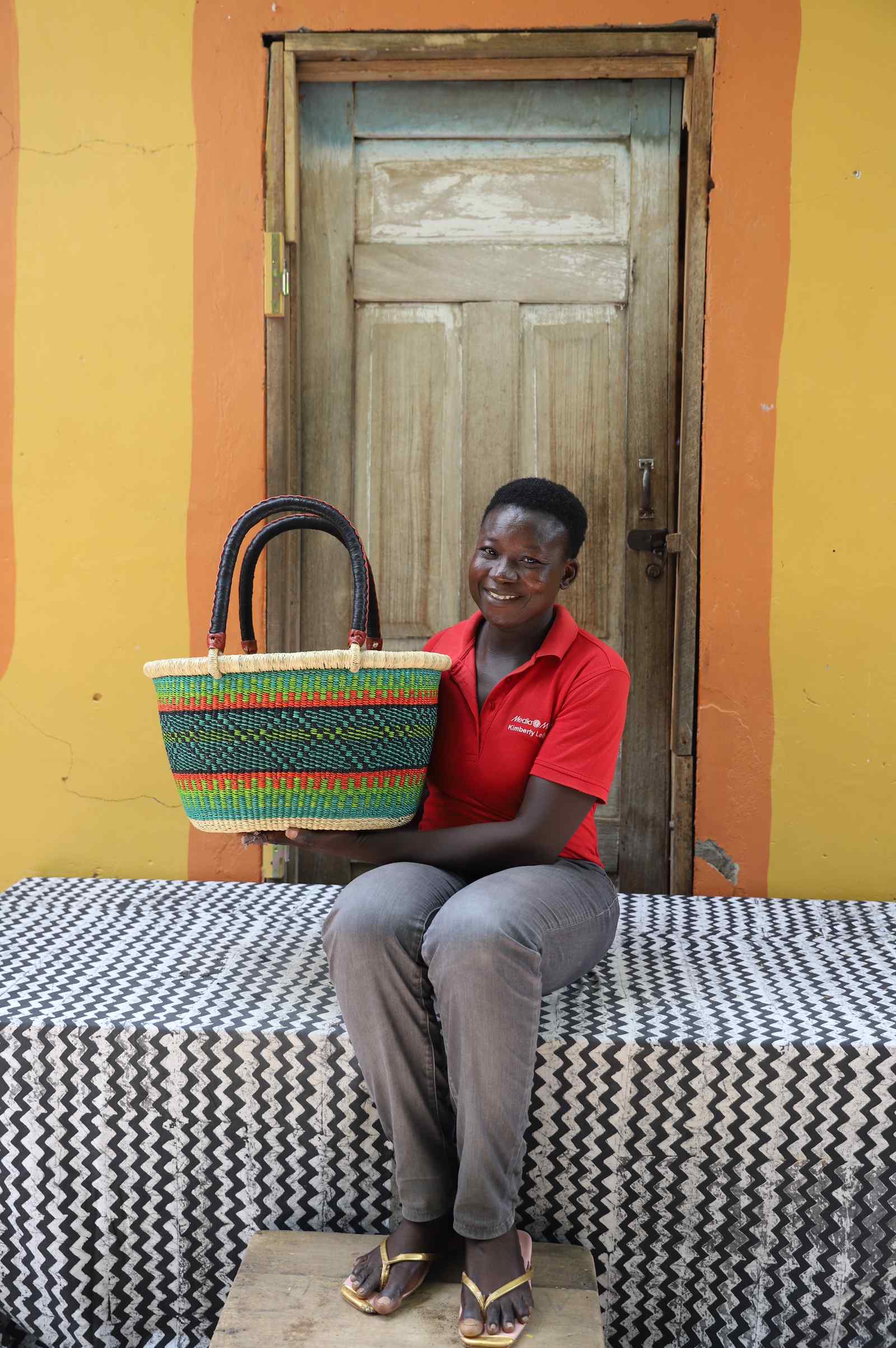 Medium oval Bolga shopping basket in green with red and yellow bands, held by a seated woman in blue and red.