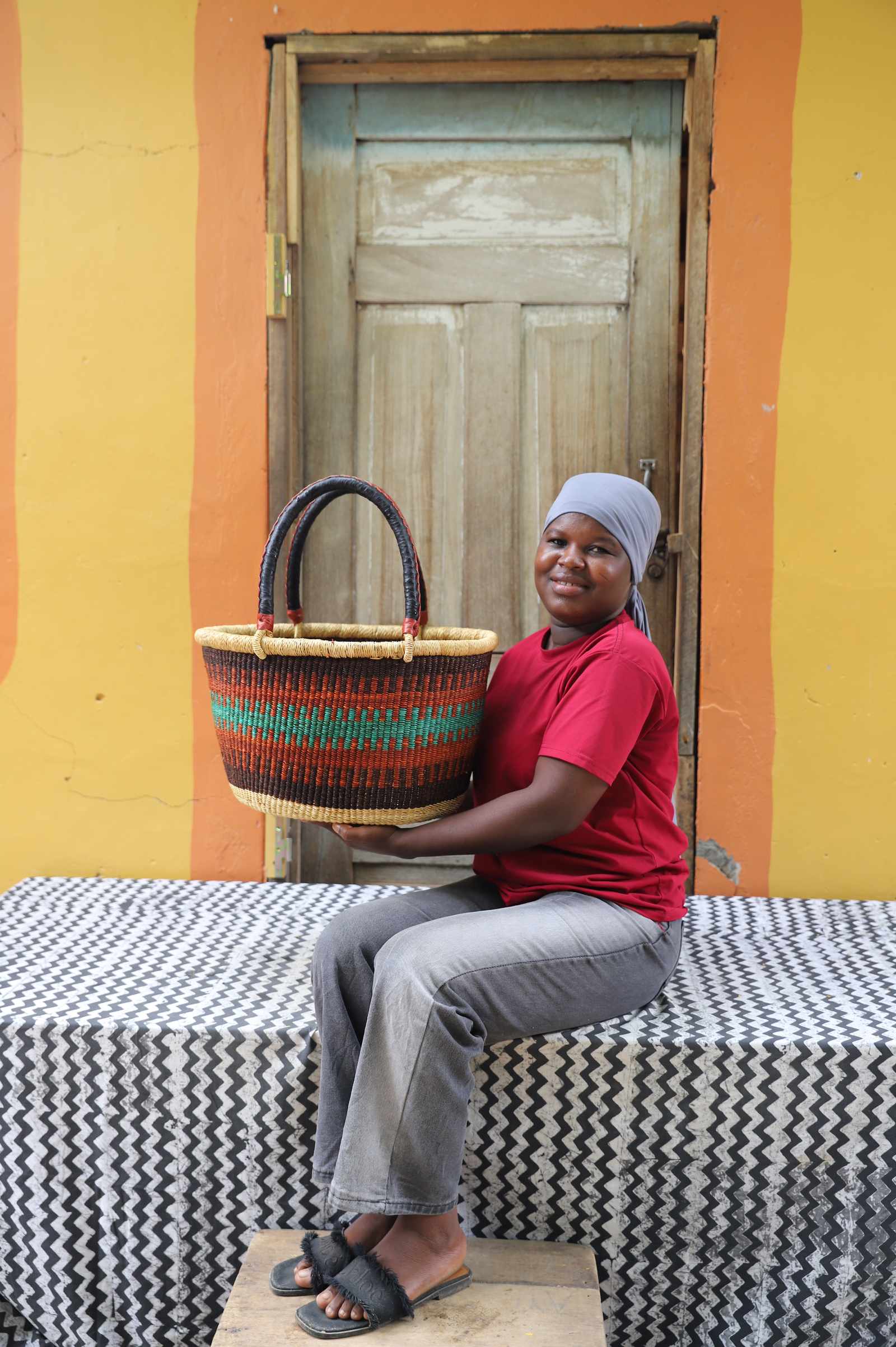 Medium oval Bolga shopping basket in natural with orange and teal bands, held by a seated woman in white.