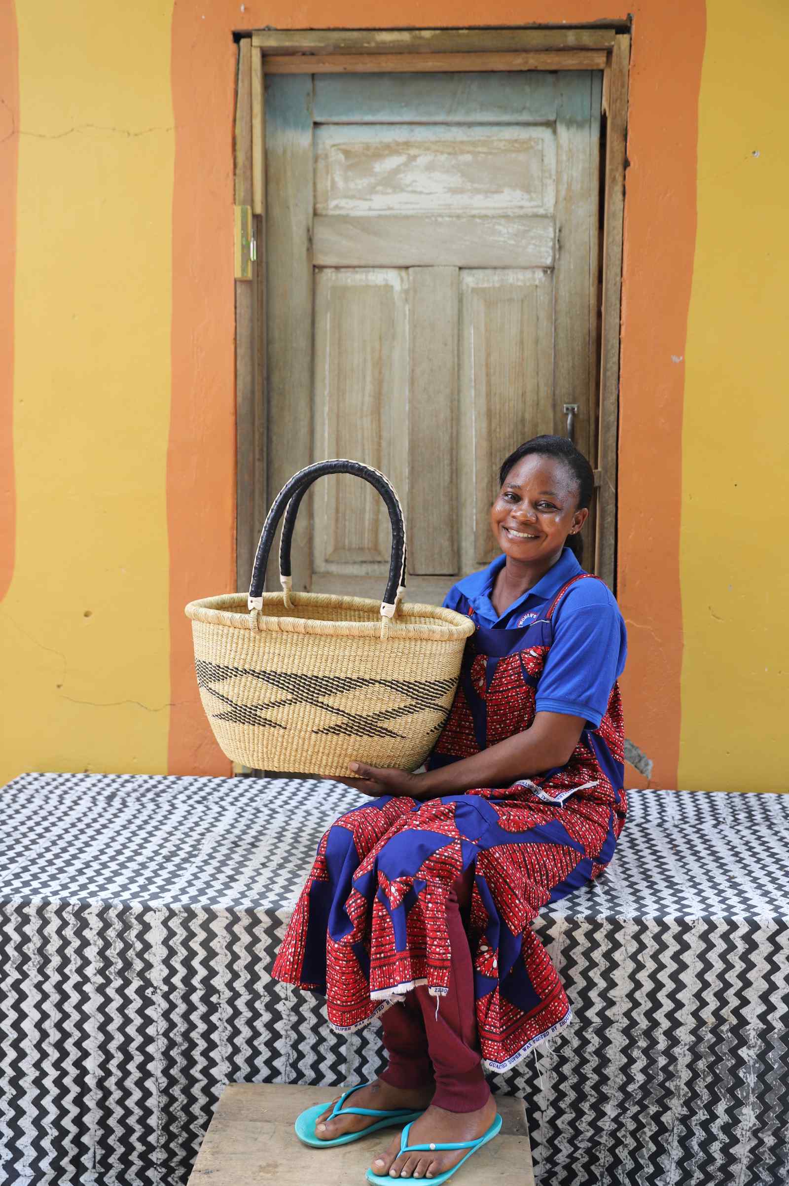Medium oval Bolga shopping basket in natural with dark bands, held by a seated woman in a printed dress.