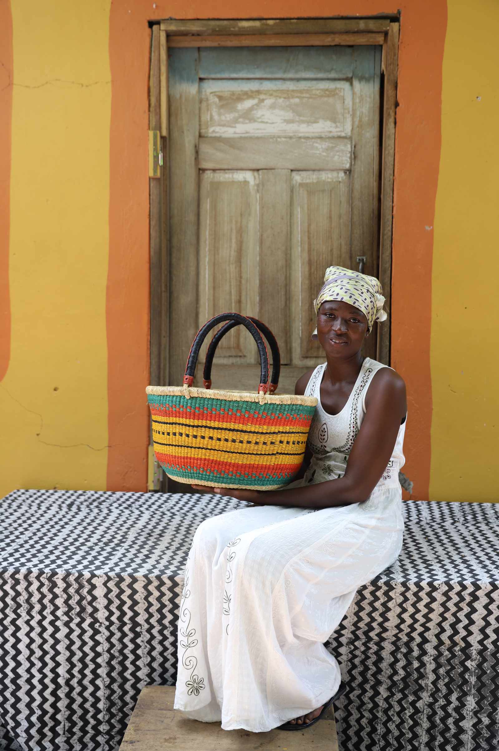 Medium oval Bolga shopping basket in natural with orange and green bands, held by a seated woman in white.