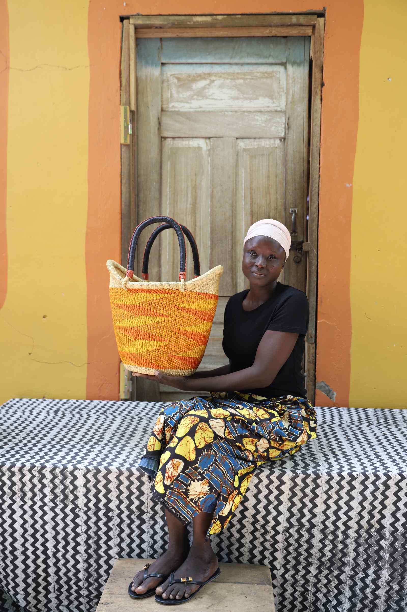 Medium Nyariga Bolga shopping basket in dyed bold orange, held by a seated woman in a floral skirt.