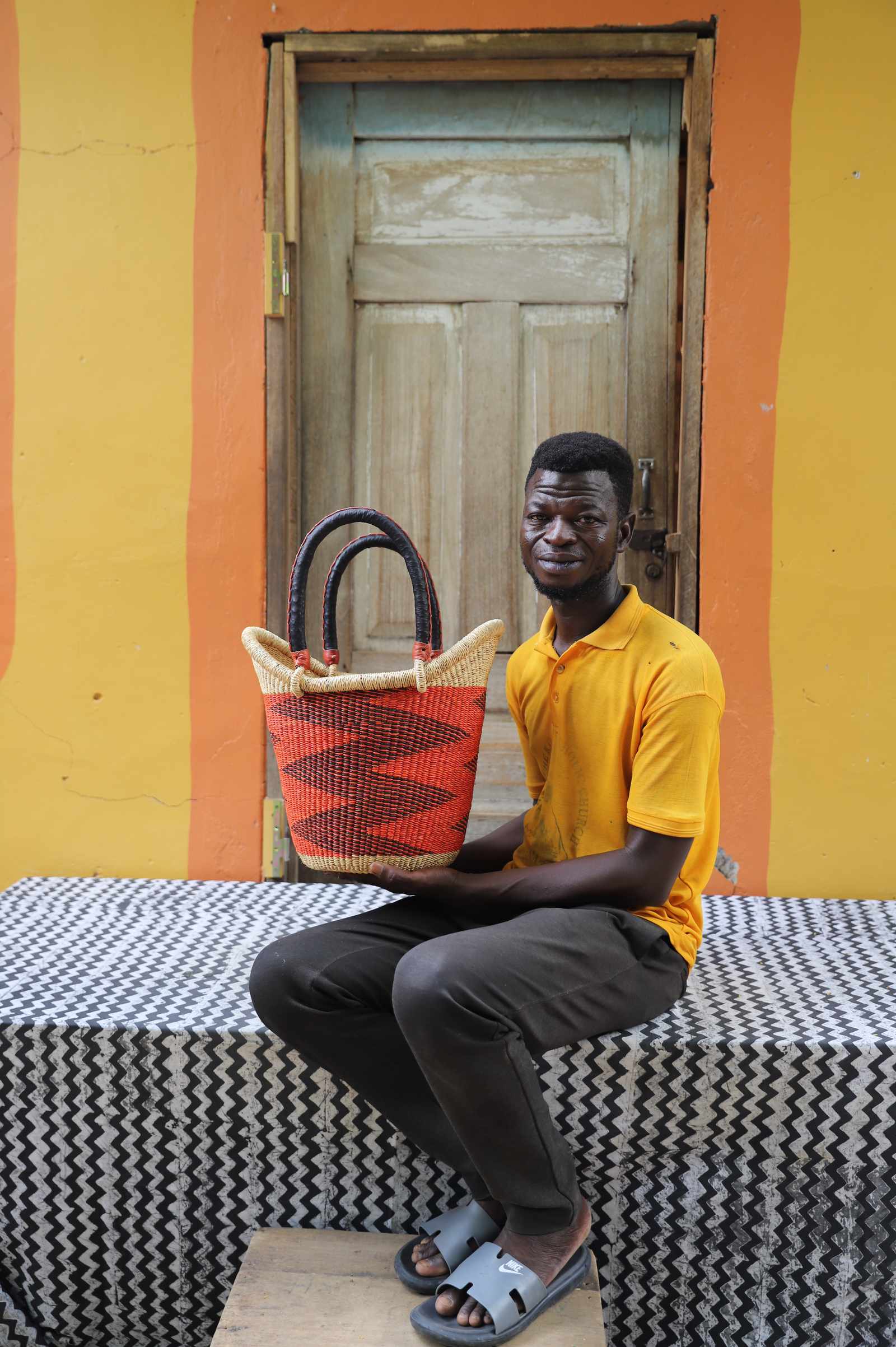 Small Nyariga Bolga shopping basket in rust-red with dark zigzag, held by a seated man in yellow.