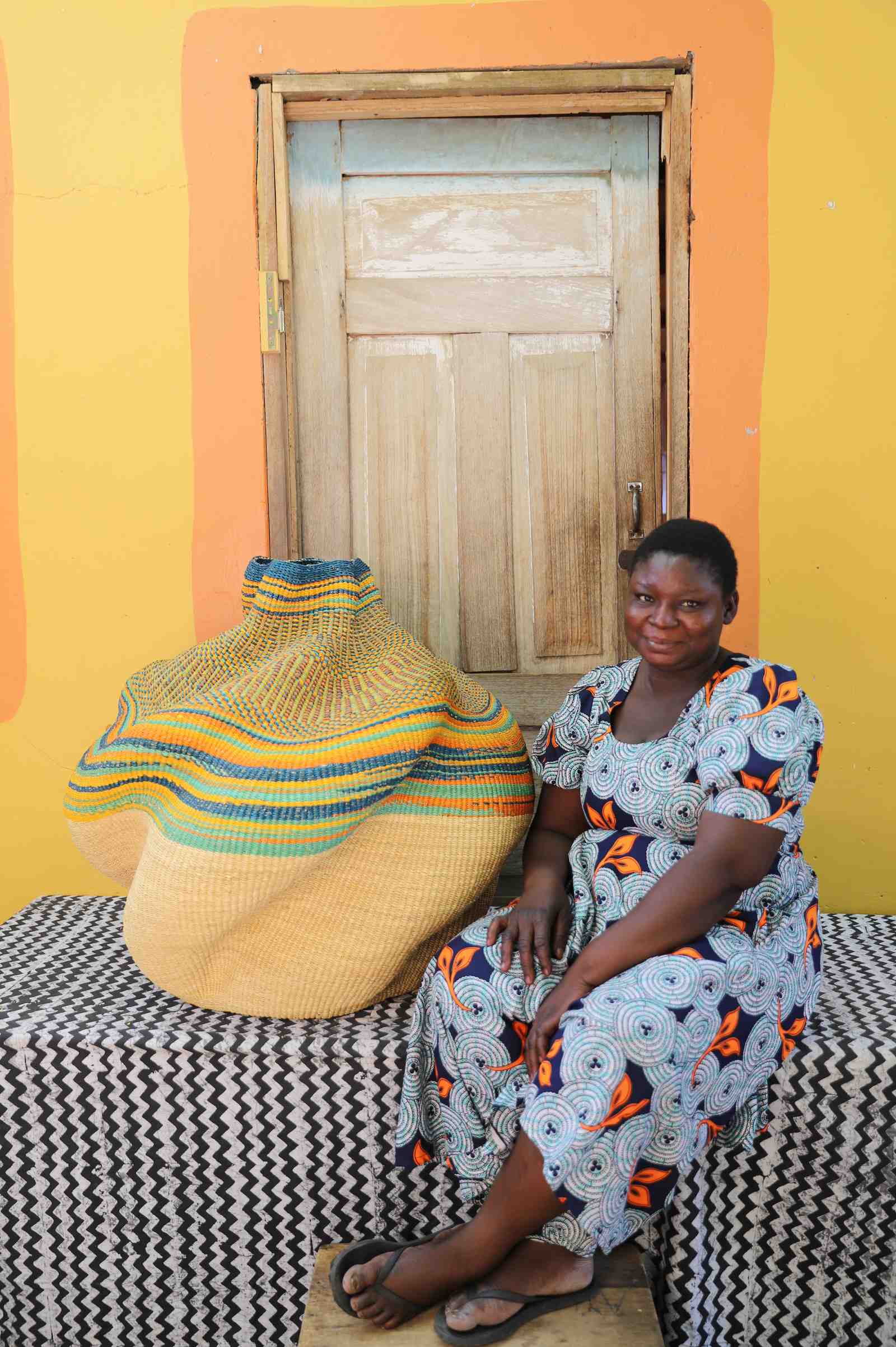Large handwoven Bolga art basket in natural straw with multicolour banded shoulder, beside a seated woman.
