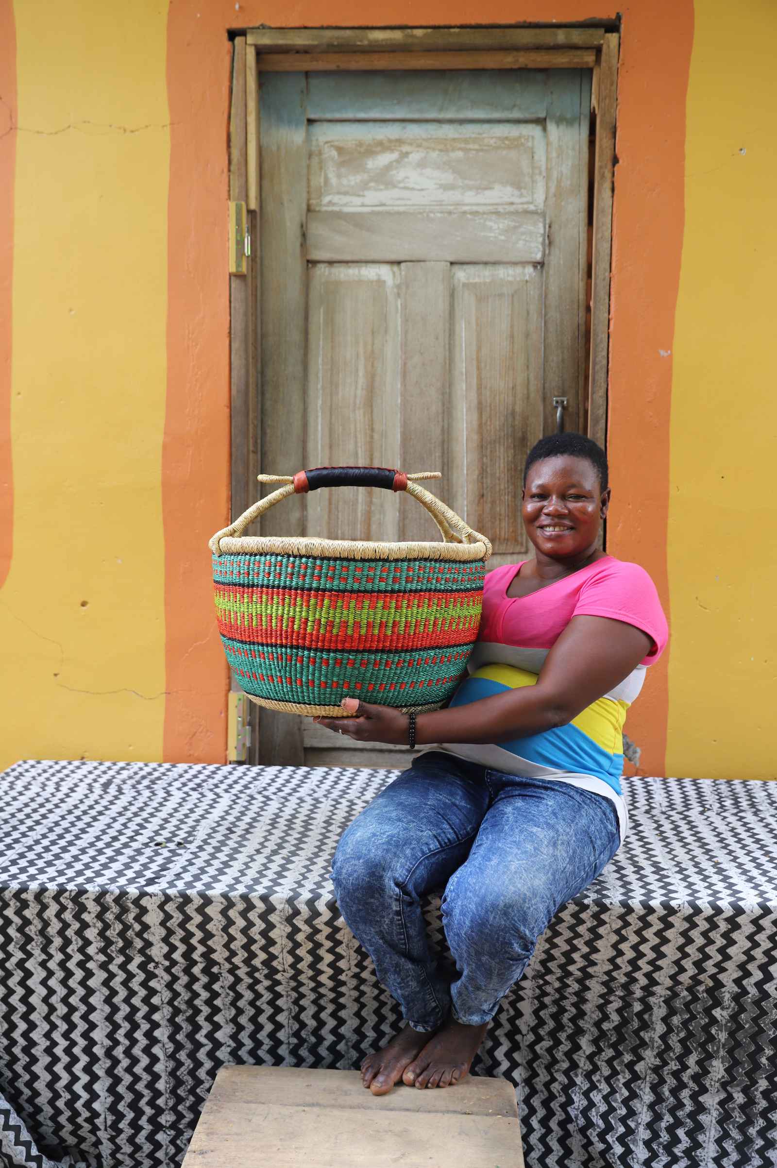 Large round Bolga shopping basket in green and red bands, held by a seated woman in pink and yellow.