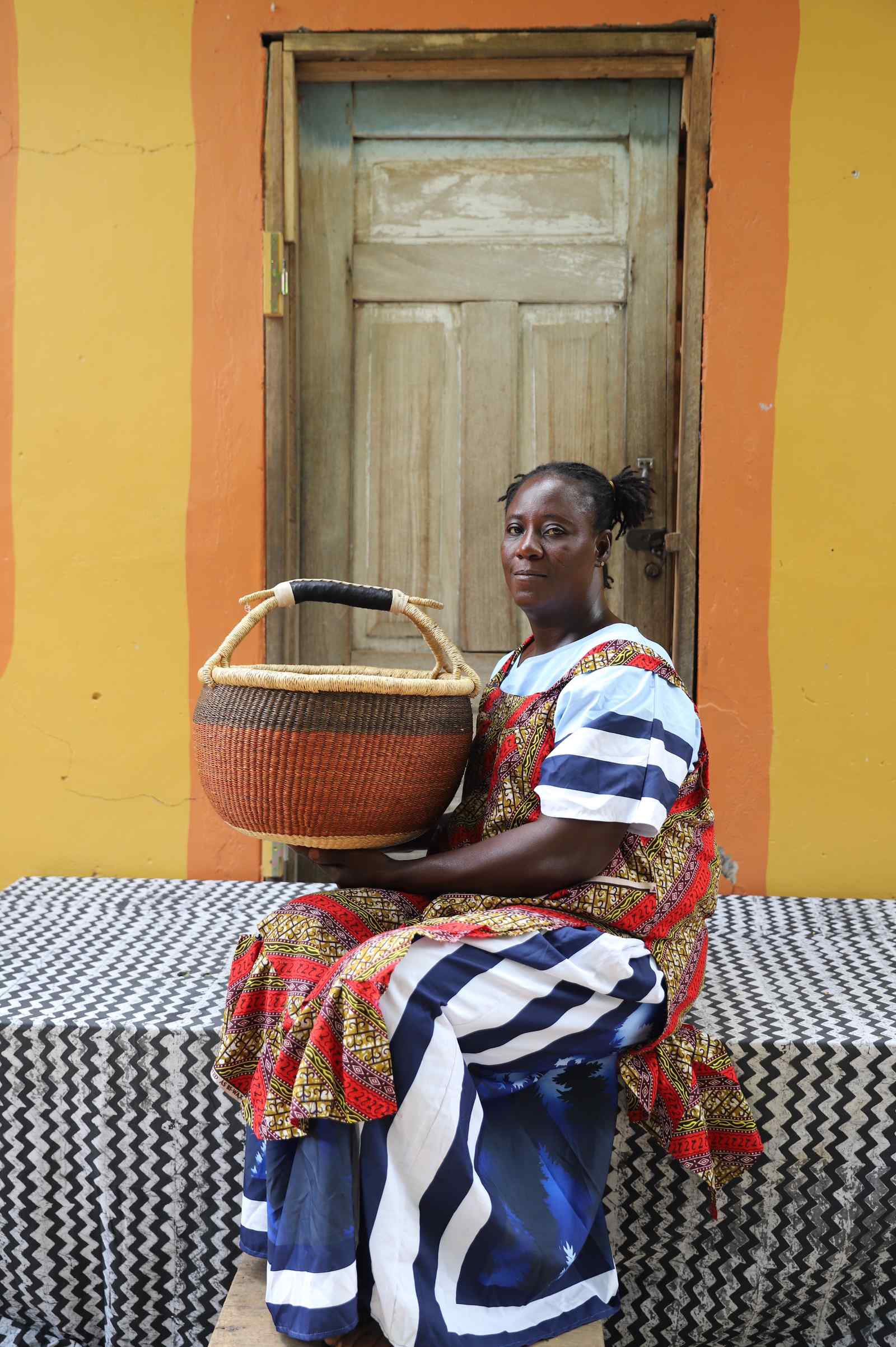 Large round Bolga shopping basket in rust-orange, held by a seated woman in a blue and white dress.