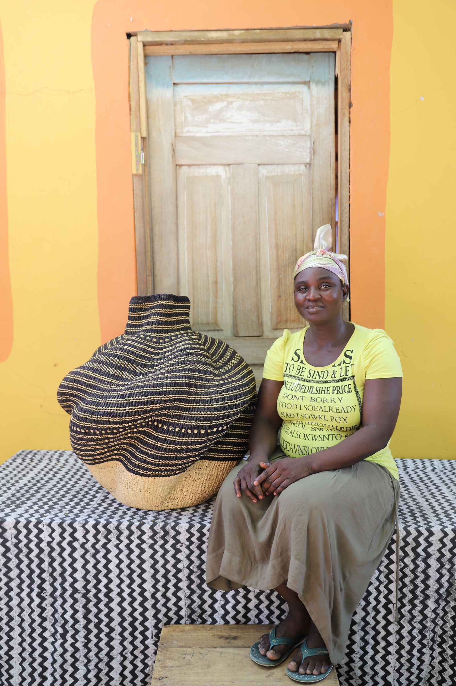 Medium handwoven Bolga art basket in natural straw with dark banded stripes, beside a seated woman in yellow.