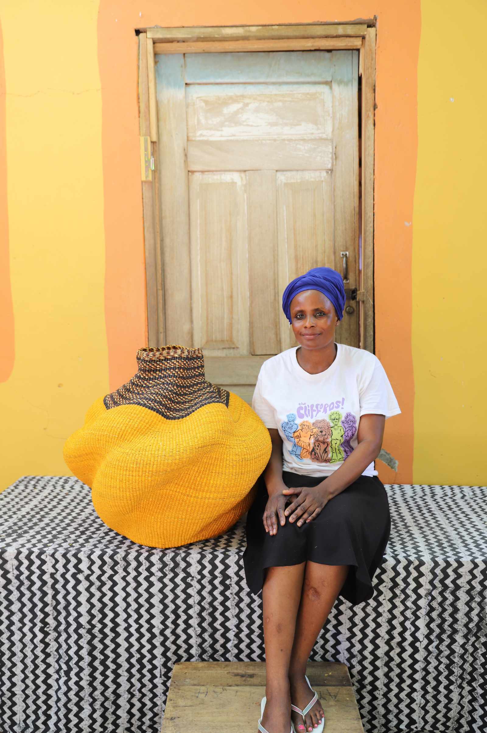 Medium handwoven Bolga art basket in mustard-yellow with a dark banded neck, beside a seated woman.