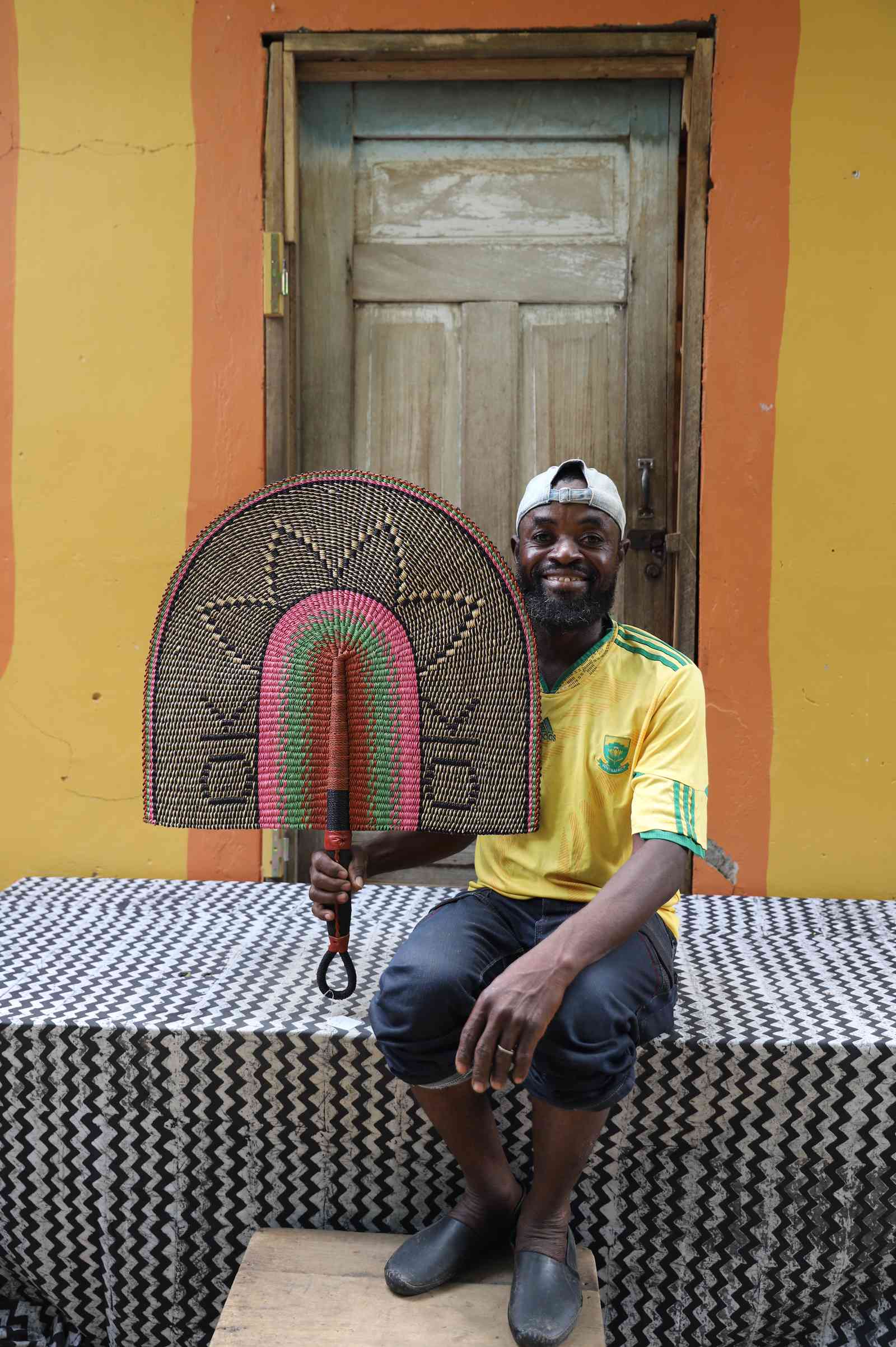 Handwoven Bolga Naba Penga wall décor fan with pink and orange floral motif on tan, held by a seated man.