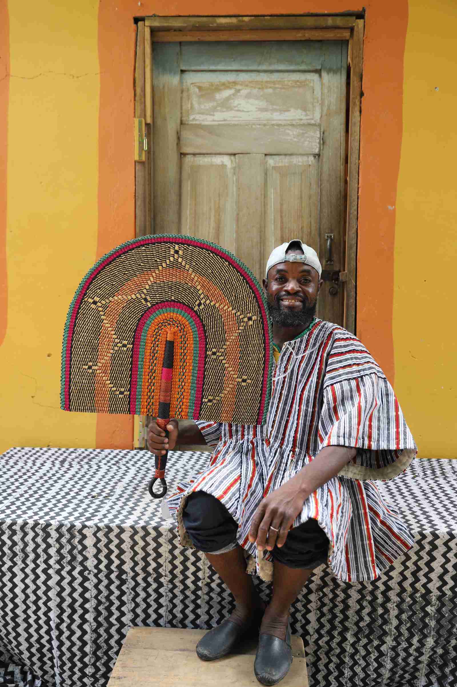 Handwoven Bolga Naba Penga wall décor fan in rust-orange arched bands with a wooden handle, held by a seated man.