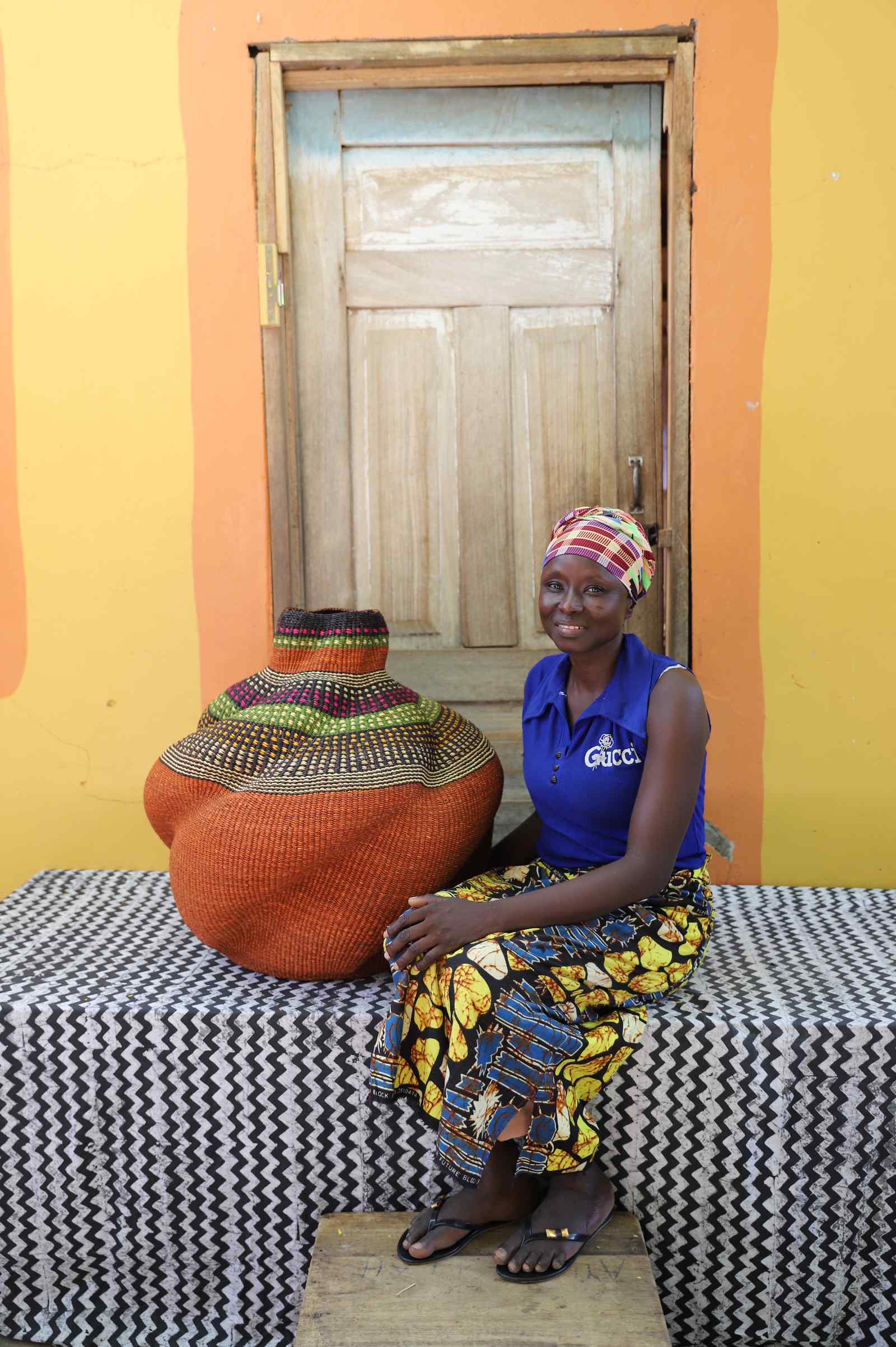 Medium handwoven Bolga art basket in rust-orange with multicolour banded shoulder, beside a seated woman.