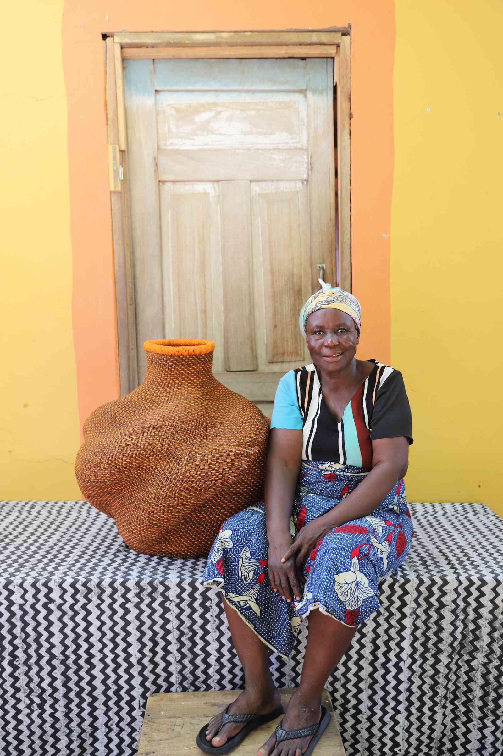 Medium handwoven Bolga art basket in rust-red with an orange rim, beside a seated woman in blue and black.